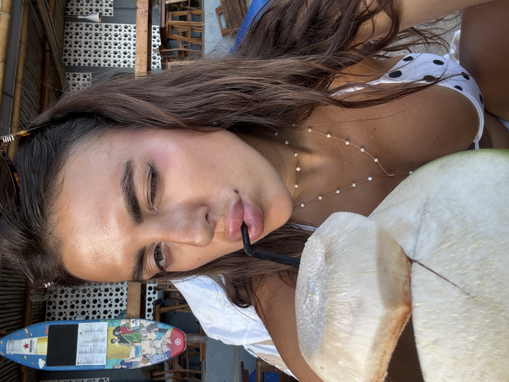 A person, likely a woman, is drinking from a young coconut with a straw. She is wearing a white top with black polka dots and a gold necklace with white beads. She is partially closing one eye. A surfboard with various images is visible below, lying flat. The setting appears to be an outdoor seating area, possibly at a beachside location, with wooden chairs and tables in the background. Based on the style of furniture and location near a beach it is probably located in Kuta Selatan, Indonesia.