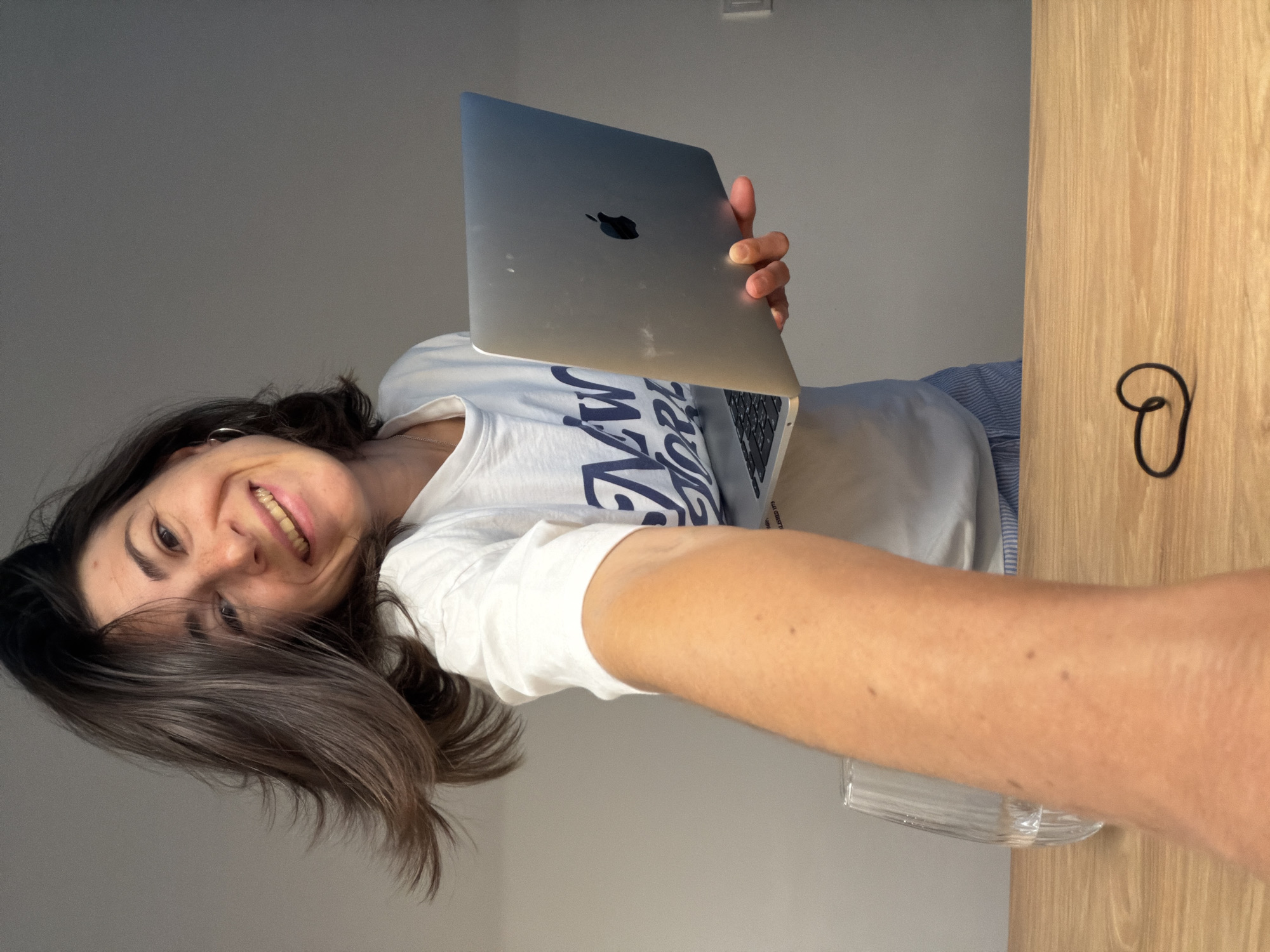A woman with dark hair and a white T-shirt is holding up a closed silver laptop with an Apple logo. She is smiling and looking at the camera. The background consists of a light grey wall and a wooden panel with a black hair tie on it. The location appears to be indoors. There is no information to indicate the image was taken in the city of Budva, Montenegro.