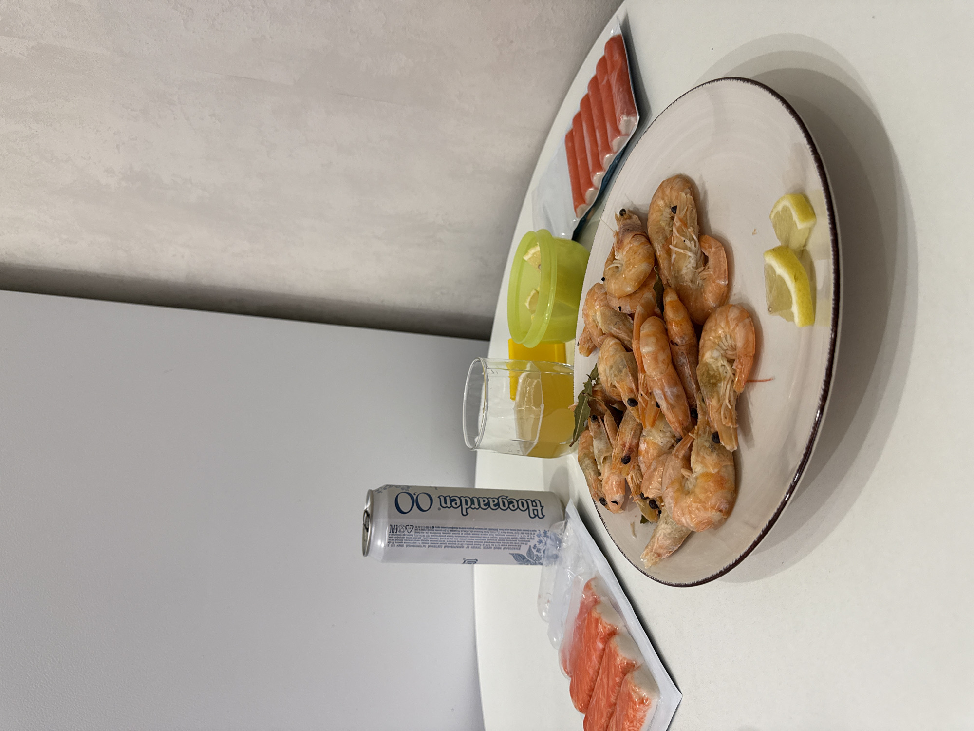 On a white table, a meal is laid out. A plate in the foreground holds a generous portion of cooked shrimp, garnished with two lemon wedges and a few bay leaves. To the left of the plate, a white plastic tray contains several rows of crab sticks, some individually packaged. Behind the shrimp plate, a tall glass of beer sits next to a small green bowl filled with lemon slices. A silver can of Hoegaarden beer stands upright next to the glass. To the right of the beer, another white plastic tray displays more crab sticks. The background consists of a plain white wall on the left and a textured, light grey wall on the right. The lighting suggests an indoor setting. The scene does not contain any individuals and there are no identifiable landmarks or specific features that would indicate a location in Vologda, Russia.