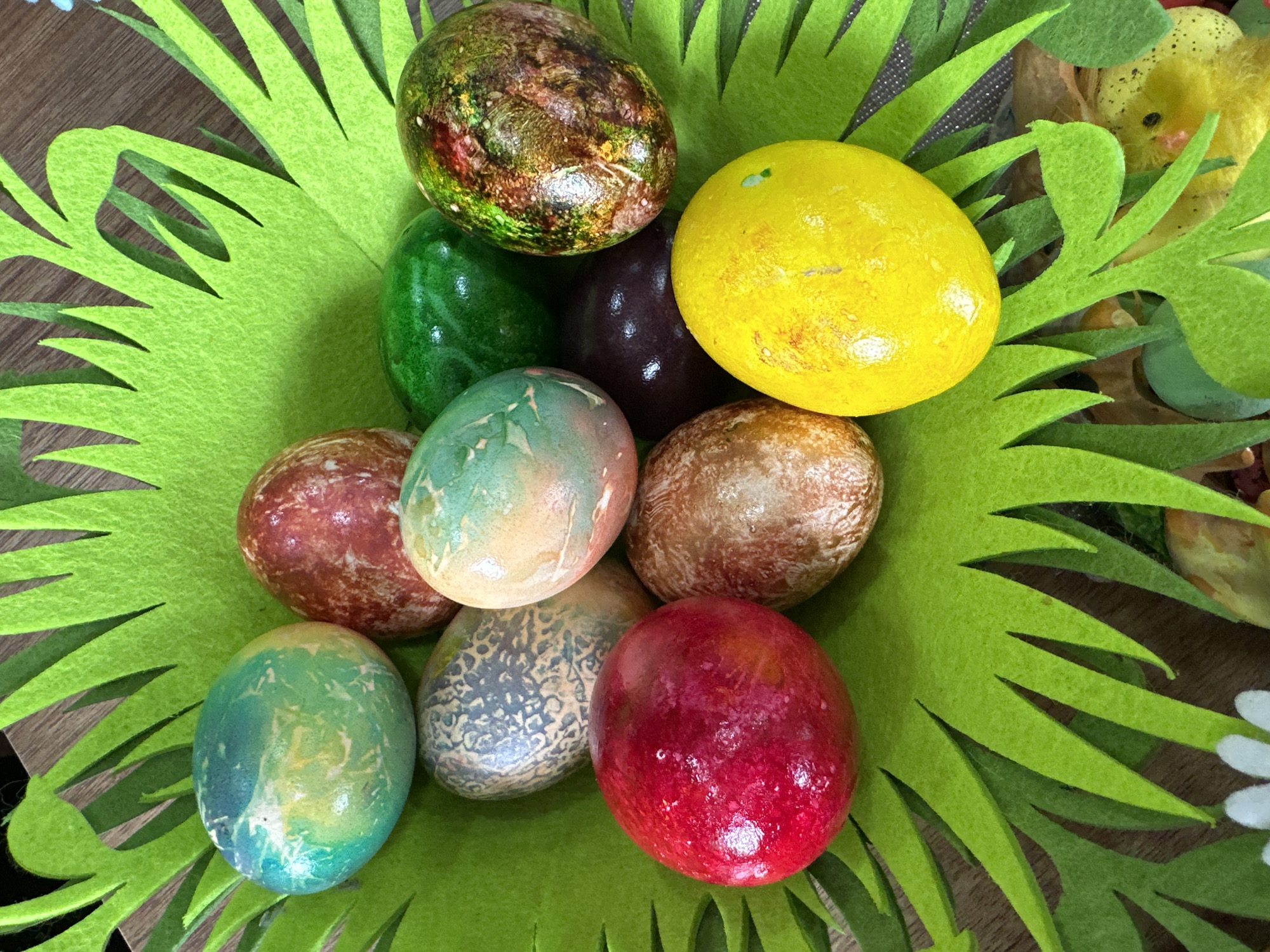 A close-up, eye-level shot shows a nest of green felt grass filled with about ten decorated Easter eggs. The eggs display a variety of colors including yellow, red, green, brown, blue, and purple, with speckled and mottled designs. A small decorative chick is nestled near the rim of the nest. The setting appears to be indoors, possibly in a home.