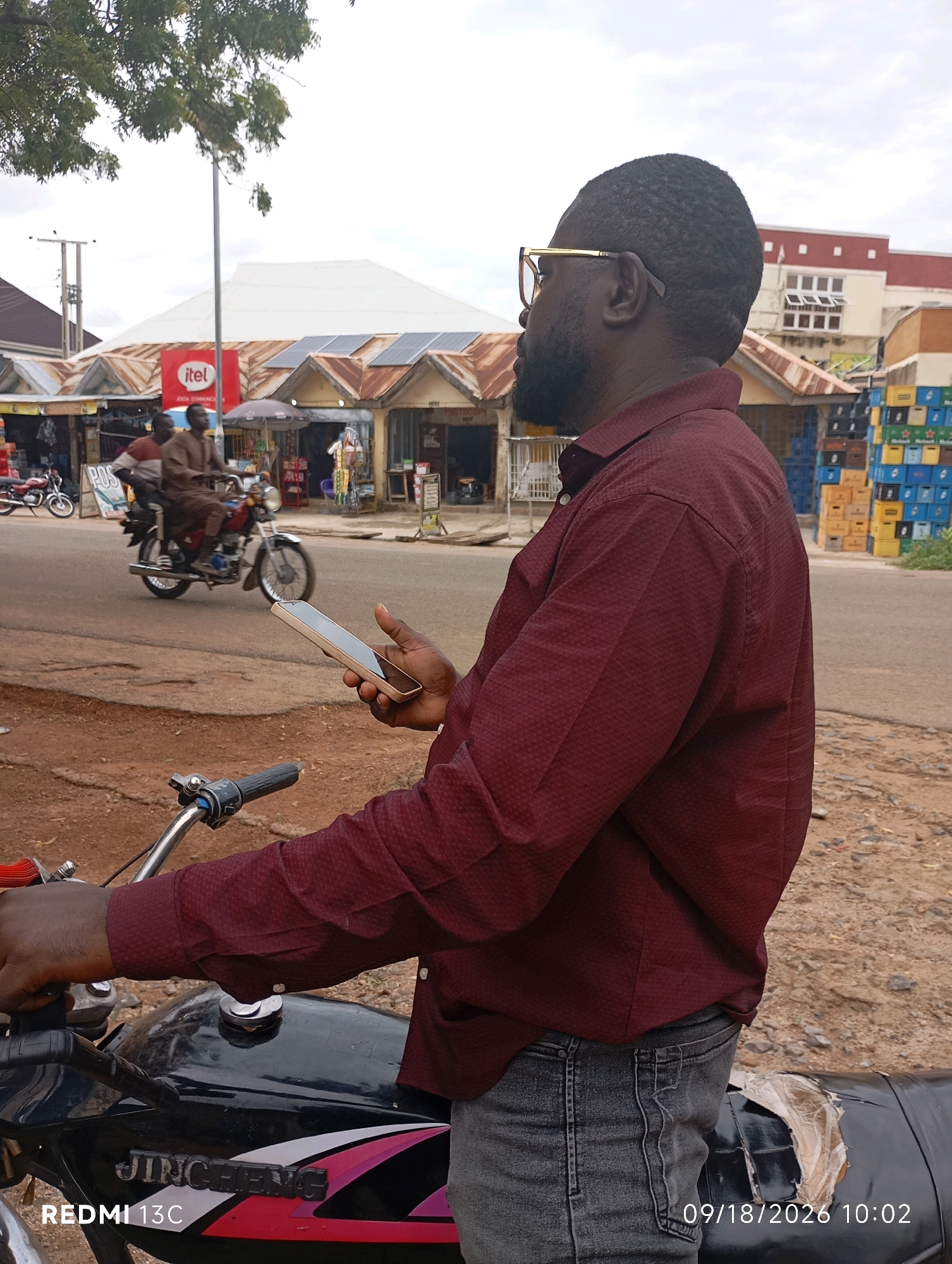 A medium shot captures a Black man, seen from the waist up and facing right, wearing a maroon patterned long-sleeved shirt and grey jeans. He is holding a gold-colored smartphone in his right hand, which is positioned in front of his chest. The man is seated on a black motorcycle with a chrome handlebar and part of the fuel tank visible. His left hand is near the handlebar.
In the background, a street scene unfolds in Shendam, Nigeria. Several small shops with corrugated iron roofs line the street. A sign for "Itel" is visible on one of the buildings, along with solar panels installed on several rooftops. Two men are riding a red motorcycle in the middle of the street, passing by the shops. Stacks of crates, some blue and yellow, are piled on the side of the road. The ground is a mixture of dirt and gravel, and the sky is overcast. The date and time stamp "09/18/2026 10:02" is visible in the lower right corner of the image, and "REDMI 13C" is in the lower left.