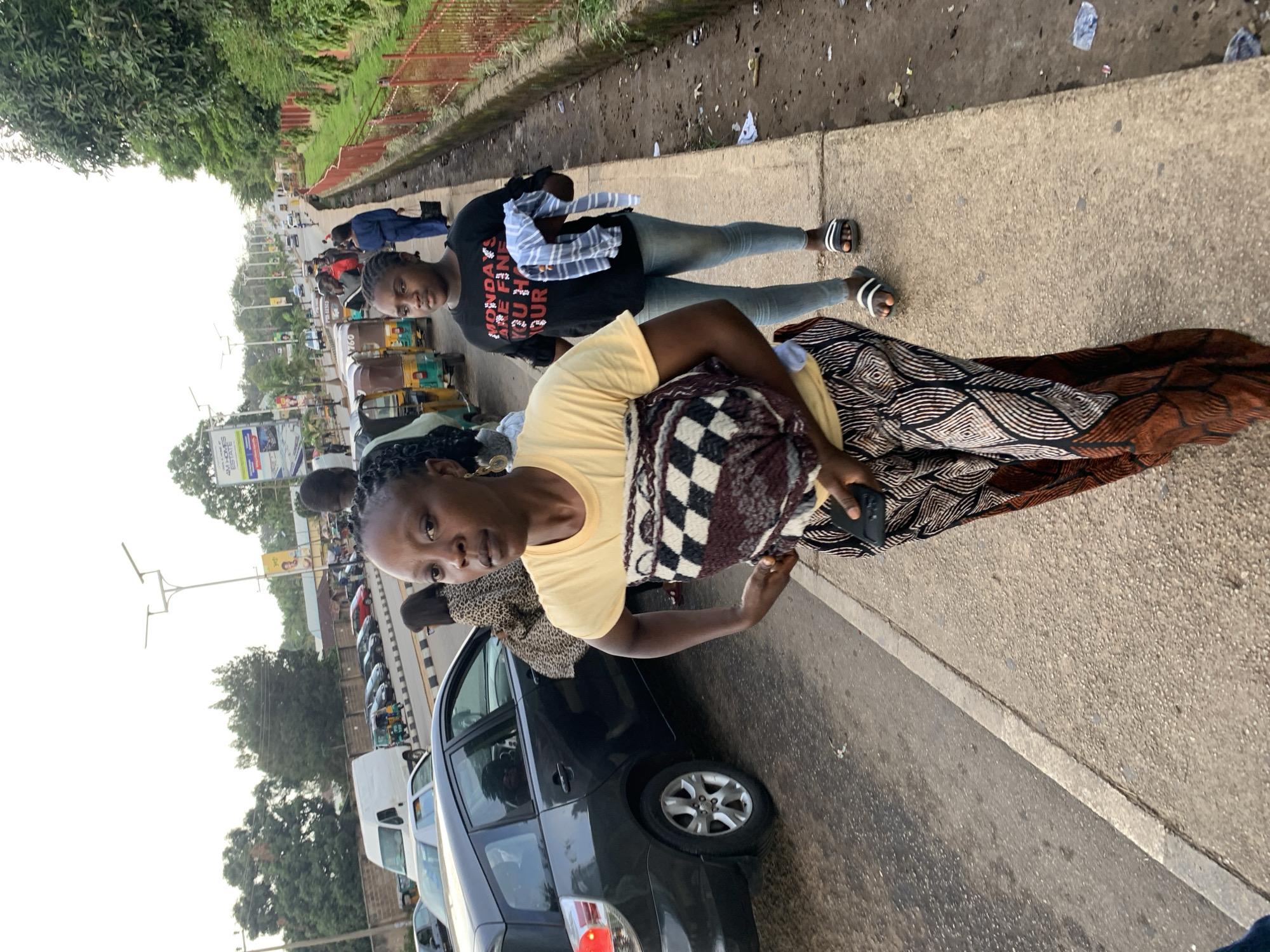 The image captures a street scene in Barakin Lerawi, Nigeria, with a focus on two women walking on a sidewalk next to a road. In the foreground, a woman with braided hair wears a yellow t-shirt and patterned trousers, carrying a patterned blanket or cloth. To her right, a younger woman in a black t-shirt and light blue jeans walks with a striped cloth in her hand. Behind them, other pedestrians, vehicles including a dark car, a white van, and auto-rickshaws, are visible on the road and sidewalk. The environment includes a red metal fence, greenery, streetlights, and signs on the side of the road, suggesting an urban or semi-urban setting. Debris is present on the edge of the sidewalk.