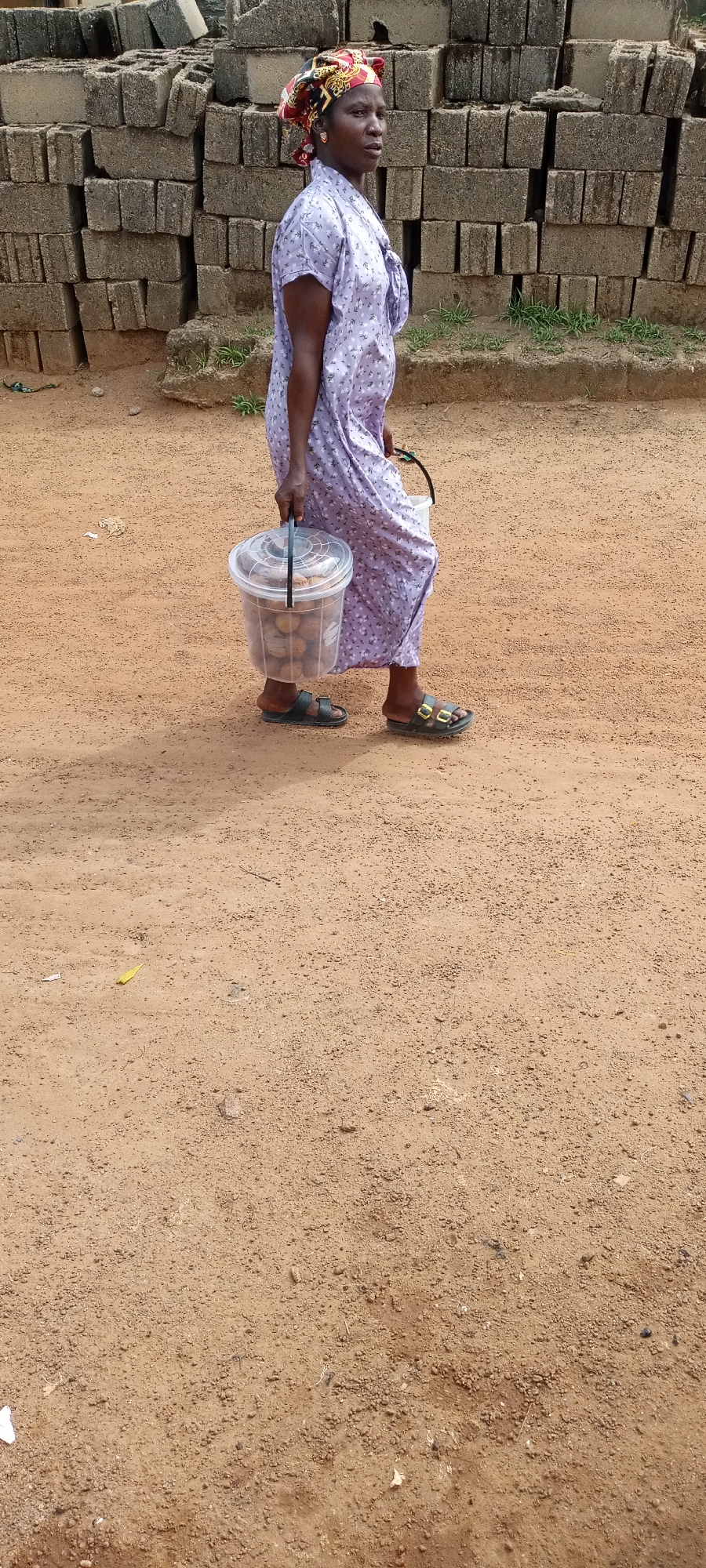 A woman wearing a purple floral dress and a colorful headscarf walks from left to right across a dirt path. She is carrying a clear plastic bucket filled with what appear to be nuts or similar small food items. In the background, a wall of grey cinder blocks is stacked high. The ground is made of brown dirt and small pebbles, with patches of dry grass. The woman is wearing dark sandals.