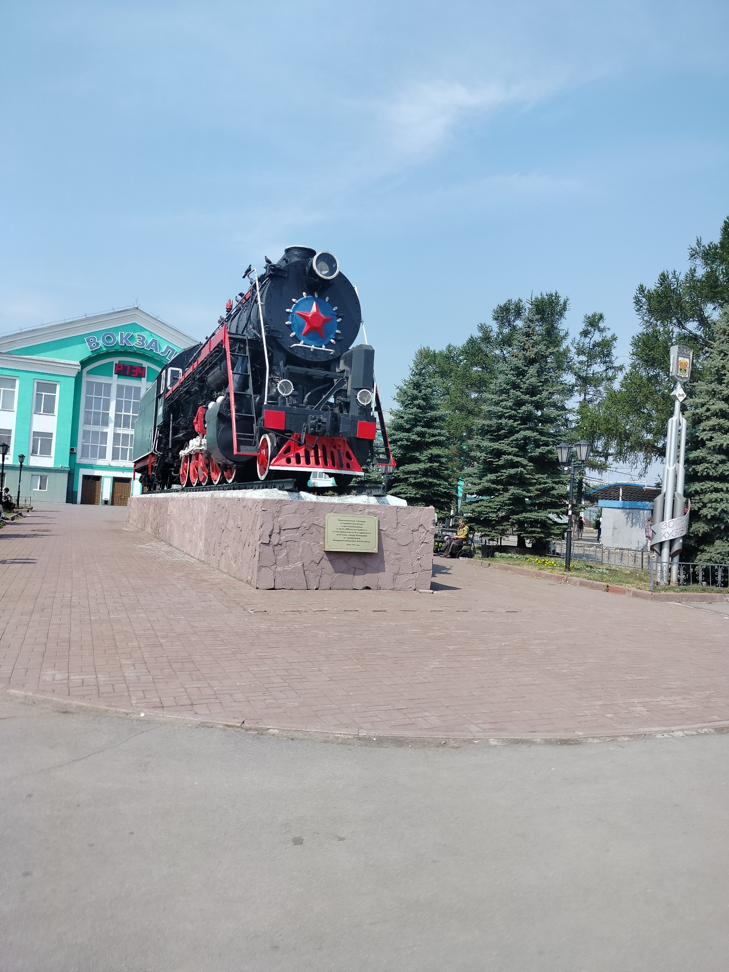 A large, black steam locomotive with red wheels and a red star emblem is on display in front of a light green building with the word "ВОКЗАЛ" (meaning "station" in Russian) in large teal letters. The locomotive sits on a stone pedestal. The scene is outdoors, with a brick-paved walkway in the foreground and a street beyond. Several evergreen trees are visible behind the locomotive. A digital clock displaying "07:34" is mounted on the building. In the background, there are streetlights and a monument. The sky is clear and blue. The location is the Kemerovo Railway Station in Kemerovo, Russia.