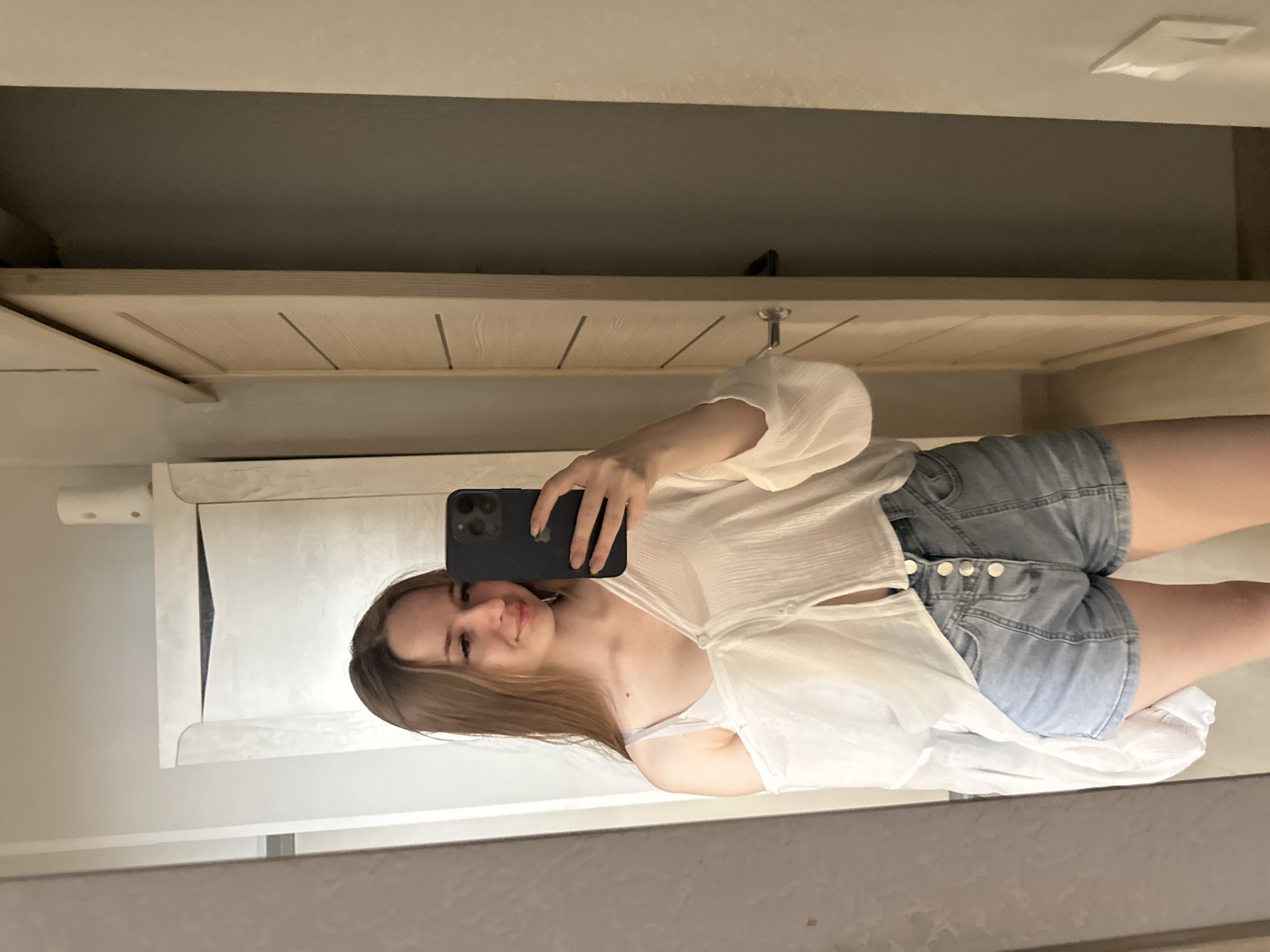 In a full-body mirror selfie, a young woman with long, light brown hair smiles as she holds a dark smartphone in her right hand to take the picture. She is wearing a white, oversized, off-the-shoulder blouse, unbuttoned at the front, revealing a white bra underneath, and light blue denim shorts with four visible buttons. She is standing in what appears to be a bedroom or dressing area, with a closet door to her right and a dresser to her left. The walls are neutral tones, and the lighting is soft.
