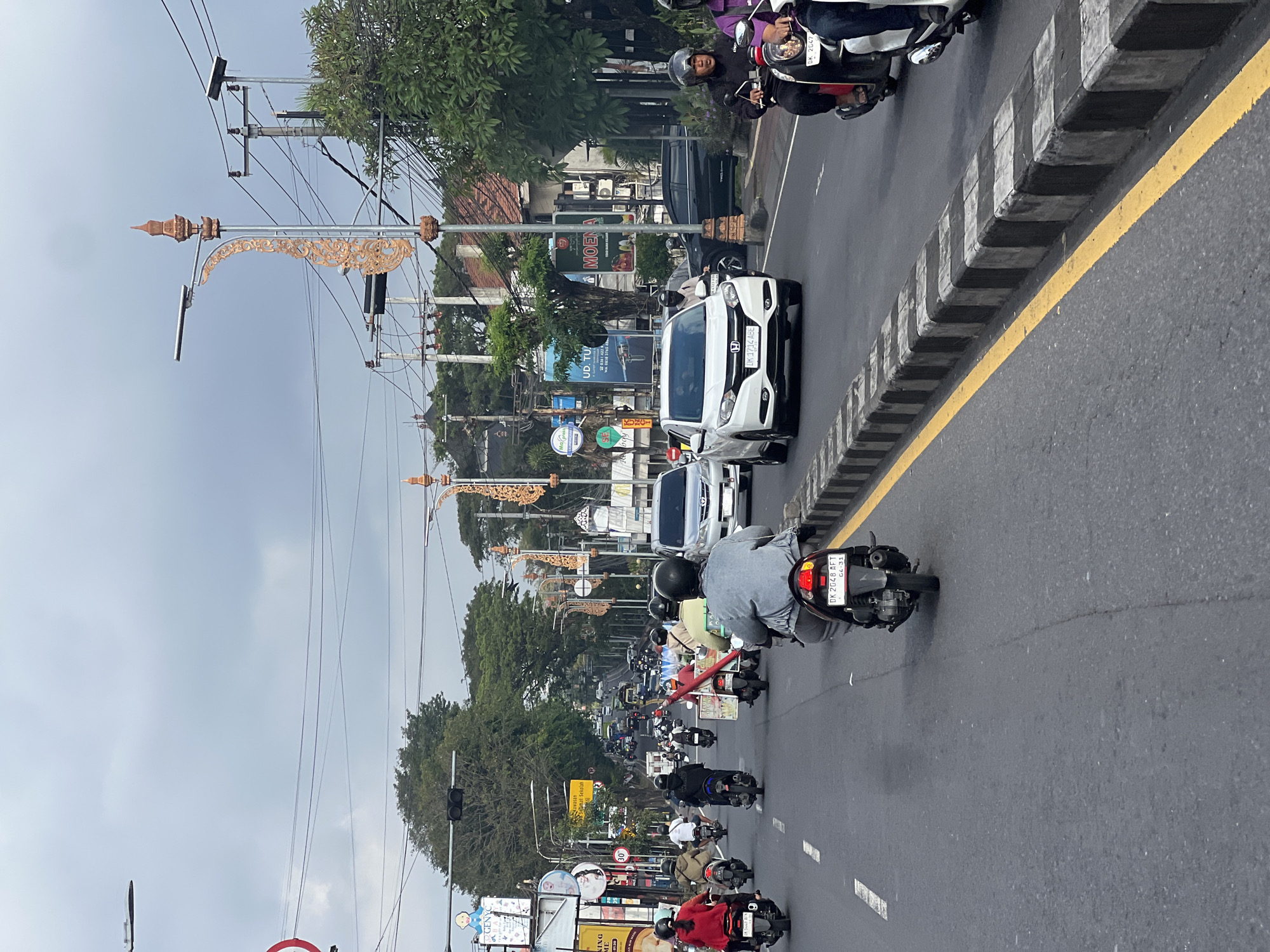 The image captures a busy street scene in Denpasar, Indonesia, on an overcast day. The perspective is from behind a motorcycle, looking forward along a wide, asphalt road. Numerous motorbikes and a few cars are navigating the traffic. Pedestrians are not clearly visible.
The roadside is lined with a raised concrete barrier, alternating black and white blocks, with a yellow line painted on the asphalt adjacent to it. On the right side of the road, there are trees and buildings, with ornate streetlights featuring decorative gold accents. Billboards and signs are visible on the left side of the road, including one with "UD. TU..." and a phone number, and another advertising "MOENA". Electrical wires are strung across the sky, supported by utility poles. The sky is cloudy, suggesting a moderate to low level of light.
The vehicles are predominantly motorbikes, with riders wearing helmets. The rider in the foreground is wearing a grey jacket and black helmet. Further ahead, a white car with license plate "DK 1714 ABE" is visible. The overall impression is of a typical urban traffic environment in Denpasar.