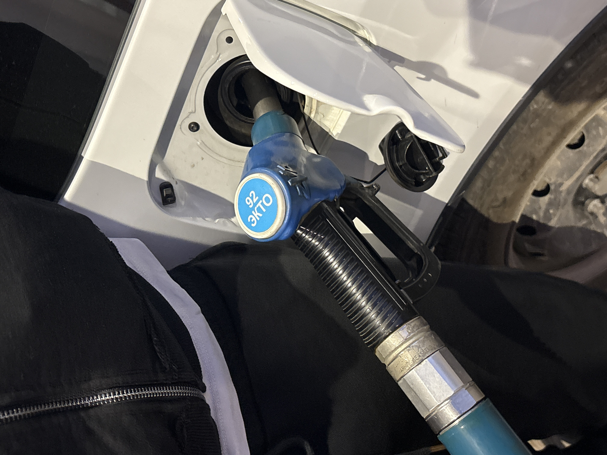 A close-up shows a blue and silver gas pump nozzle inserted into the fuel tank of a white vehicle. The vehicle's fuel door is open, revealing the black fuel tank opening. A tire is visible on the right side of the frame. The person filling the gas tank is wearing black clothing with a white stripe, and only their clothing is visible. The nozzle has a sticker indicating '92 ЭКТО', suggesting the octane rating of the fuel. The scene is likely a gas station in Perm, Russia, due to the Russian language on the nozzle.
