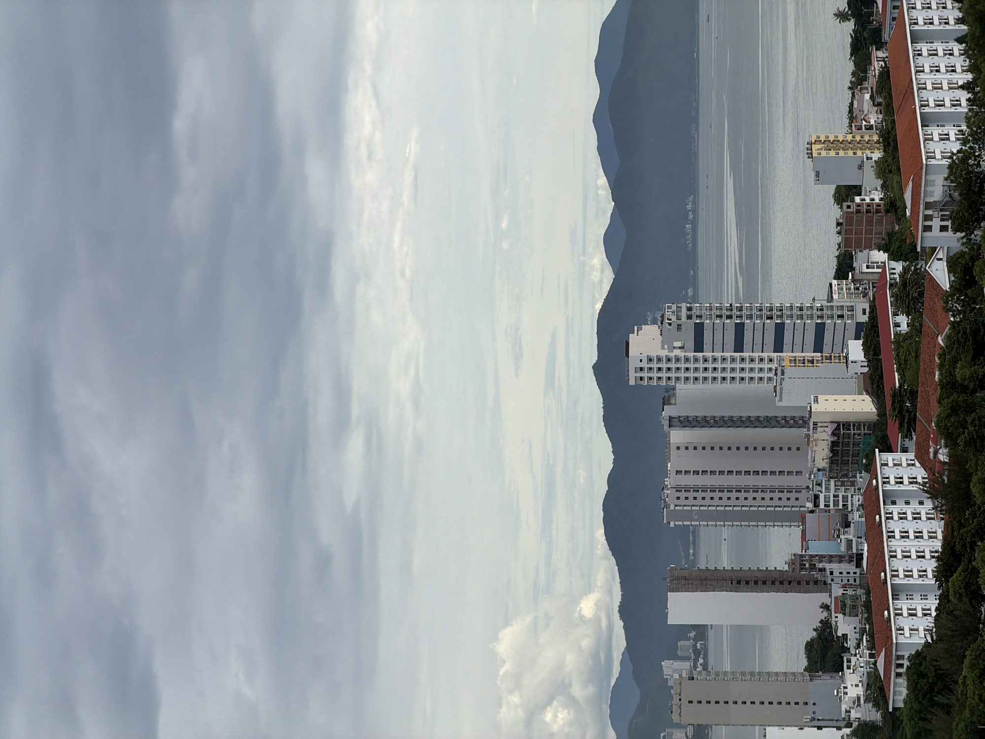 The image depicts an urban skyline in Nha Trang, Vietnam, characterized by a vast expanse of overcast sky dominating the upper portion. Below, a body of water, likely the sea, stretches towards a range of dark, undulating mountains in the distance. The middle ground is filled with a dense collection of buildings, predominantly tall, modern high-rise apartments and hotels. These structures vary in height and architectural style, with many featuring balconies and repeating window patterns. Some buildings appear to be under construction, indicated by exposed frameworks. Closer to the foreground, lower-rise residential buildings with red-tiled roofs are visible, interspersed with lush green trees. The overall scene suggests a coastal city with a mix of residential and commercial development, set against a backdrop of natural mountainous terrain. There are no discernible people or specific actions taking place.