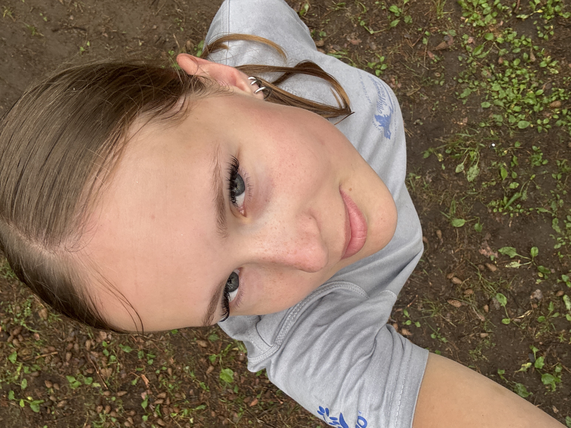 This is a close-up, high-angle selfie of a young person outdoors. The subject has light skin with freckles on their nose and cheeks, and light brown hair that is pulled back. They are wearing a light grey long-sleeved shirt with a blue graphic on the left sleeve. The person is looking up towards the camera with a slight smile. Their left ear is visible, adorned with a silver spiral earring.
The background consists of dark, damp soil with scattered green plants and some small brown debris, suggesting a natural, possibly wooded or park-like setting. The lighting appears to be natural daylight, but the overcast conditions or shadows may indicate a cloudy day or the subject is in a shaded area. The ground texture and presence of vegetation are the primary environmental cues. No specific time of day or weather beyond potentially overcast conditions can be definitively determined.
Visible text on the shirt is in Russian and appears to be a logo or team name, with the words "Чайковский" (Chaikovsky) clearly legible, indicating the location as Chaikovsky, Russia. The graphic on the sleeve features a bird in flight. The overall impression is a casual, candid moment captured outdoors.