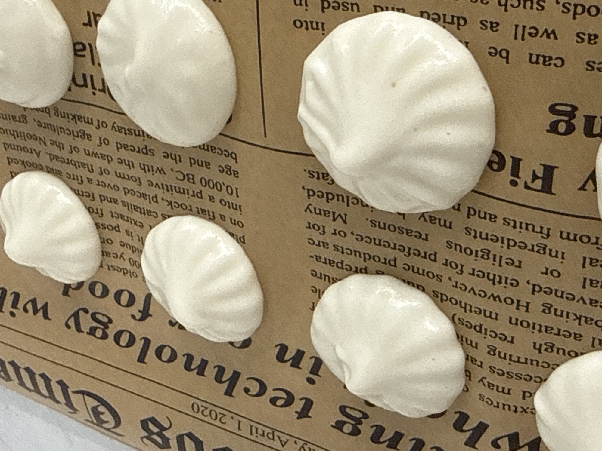The image displays a close-up view of several small, white, irregularly shaped meringue cookies placed on a textured brown surface resembling newsprint. The cookies are positioned at various angles, with some overlapping and others casting subtle shadows. The background surface has visible text in a dark font, suggesting it is a printed material. The lighting appears to be soft, highlighting the glossy texture of the meringue. There is no visible indication of the city of Lyubertsy, Russia, or any elements that would place the scene in a specific urban or geographical location. The focus is entirely on the arrangement and texture of the meringue cookies against the patterned background.
