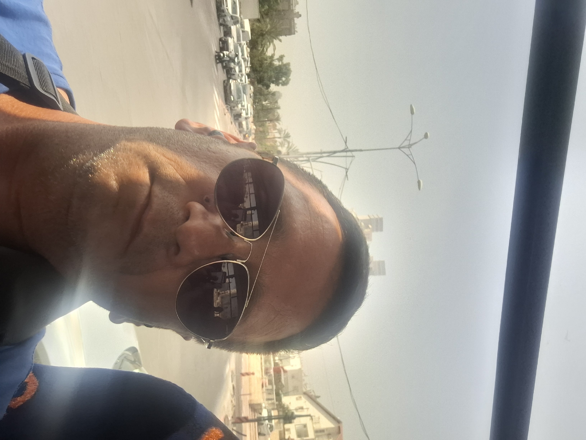 The image is a selfie taken by a man wearing sunglasses and a blue shirt, possibly on a bus or vehicle. In the background, a street scene in Be'er Sheva, Israel, is visible, featuring a traffic jam with cars and motorcycles. Palm trees and buildings line the street, and a street lamp is present. The overall atmosphere appears to be hazy and sunny, consistent with an urban environment.