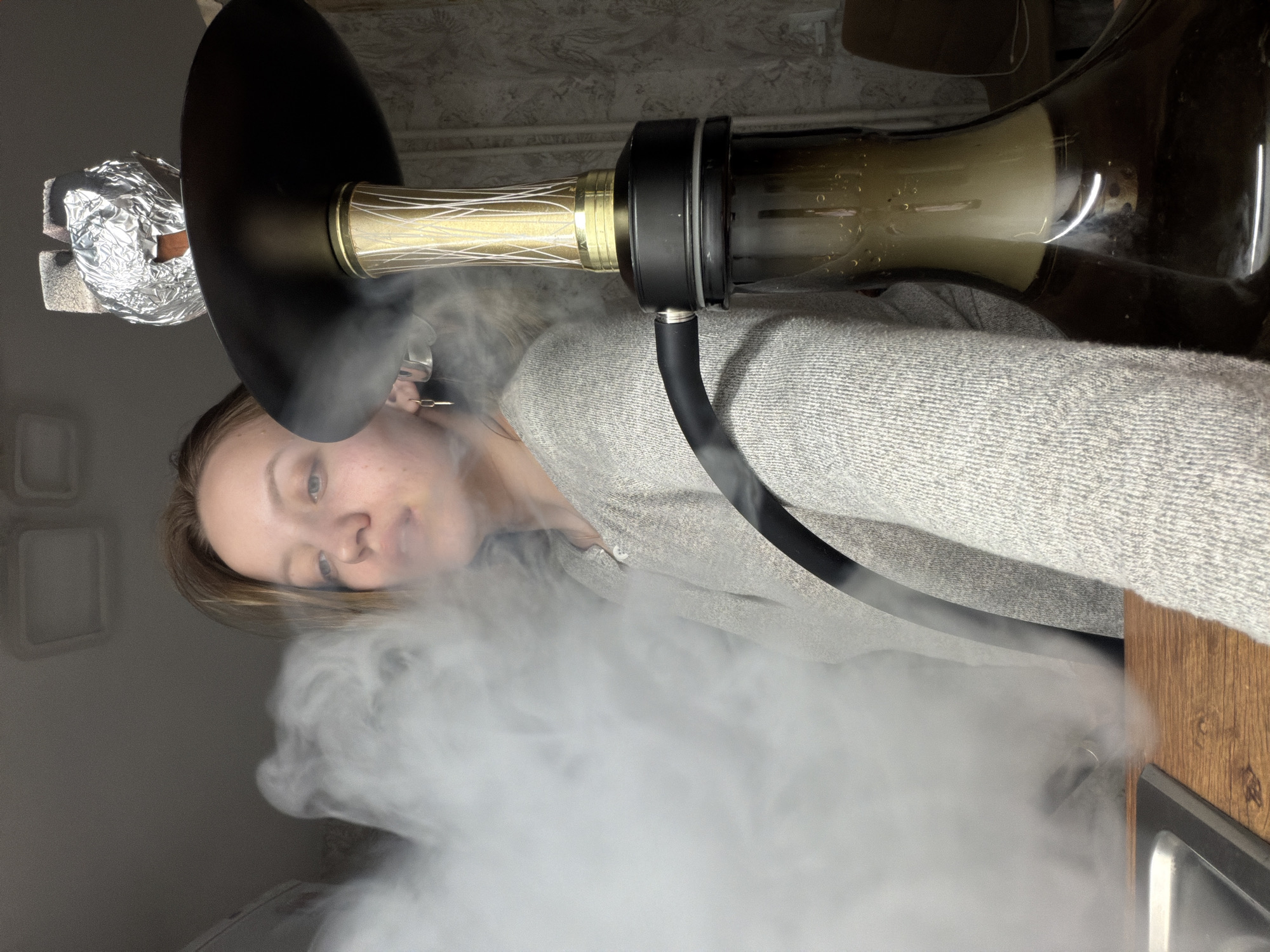 A woman is shown in a close-up shot with a hookah. The hookah is emitting a large plume of white smoke, which partially obscures the woman. She is wearing a light gray sweater. The hookah itself has a gold and black stem and a brown glass base. The hookah is set on a wooden surface next to a metal sink. The background is a neutral-colored wall. The location is not identifiable as Reshetnikovo, Russia, from the details in the image.