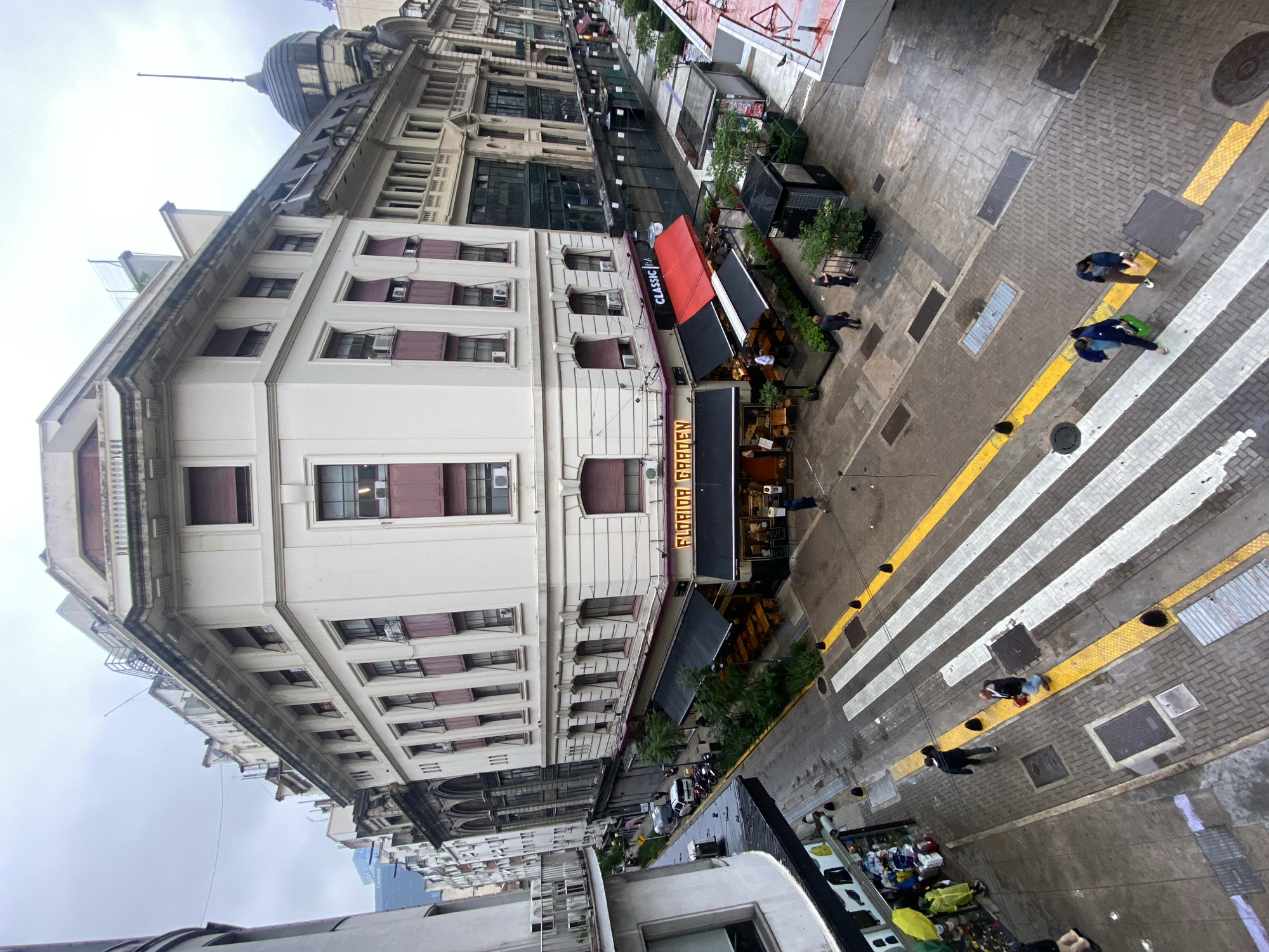 This elevated, tilted photograph presents an urban street scene in Buenos Aires, Argentina, under an overcast sky. The perspective looks down from a building, capturing a multi-story, ornate edifice on the left and the street life below.
The dominant building on the left is a grand, light-colored structure featuring intricate architectural details, multiple floors, and windows with distinctive maroon shutters or awnings. Several air conditioning units are visible beneath these windows. Further along the street, the dark dome of another stately building is visible against the cloudy sky.
At street level, a commercial establishment, likely a bar or restaurant, stands out with a prominent red awning displaying "CLASSIC BAR" in white lettering. Below it, a vertical sign reads "FLORIDA GARDEN". This venue extends onto the sidewalk with an outdoor seating area shaded by dark awnings and adorned with potted plants, where a few people are seated or standing.
The street is paved with interlocking blocks and features a well-defined pedestrian crosswalk with wide white stripes bordered by yellow lines. Several pedestrians traverse the street and sidewalks. One individual is notably seen pulling a bright green suitcase across the crosswalk. Manhole covers and grates are visible on the pavement, which appears slightly damp in places, suggesting recent rain or general moisture from the overcast conditions.
The soft, diffused lighting indicates an overcast day. The architecture and the name "FLORIDA GARDEN" are strong cues to the location being Buenos Aires. The scene captures a typical daytime urban environment with ongoing pedestrian activity.
