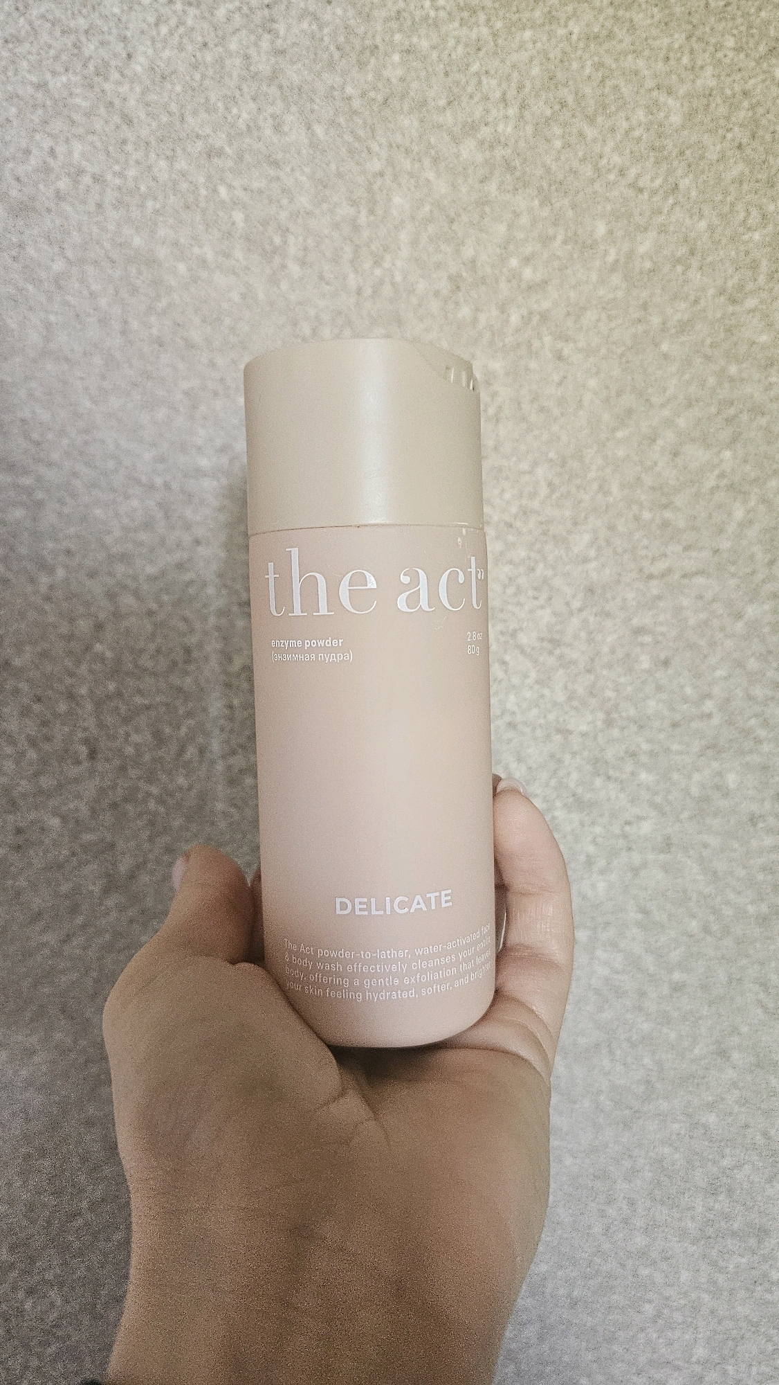 A hand holds a pale pink bottle of "the act" enzyme powder cleanser. The bottle has a beige cap and text printed in a light silver font. The text on the bottle reads "the act" at the top, followed by "enzyme powder (энзимная пудра)", and then "2.8 oz 80g". Below this, the word "DELICATE" is printed in larger letters. A description of the product is also visible in smaller text at the bottom of the bottle. The background is a textured, light grey wall. The image does not contain any elements that suggest a location in Rostov-on-Don, Russia.