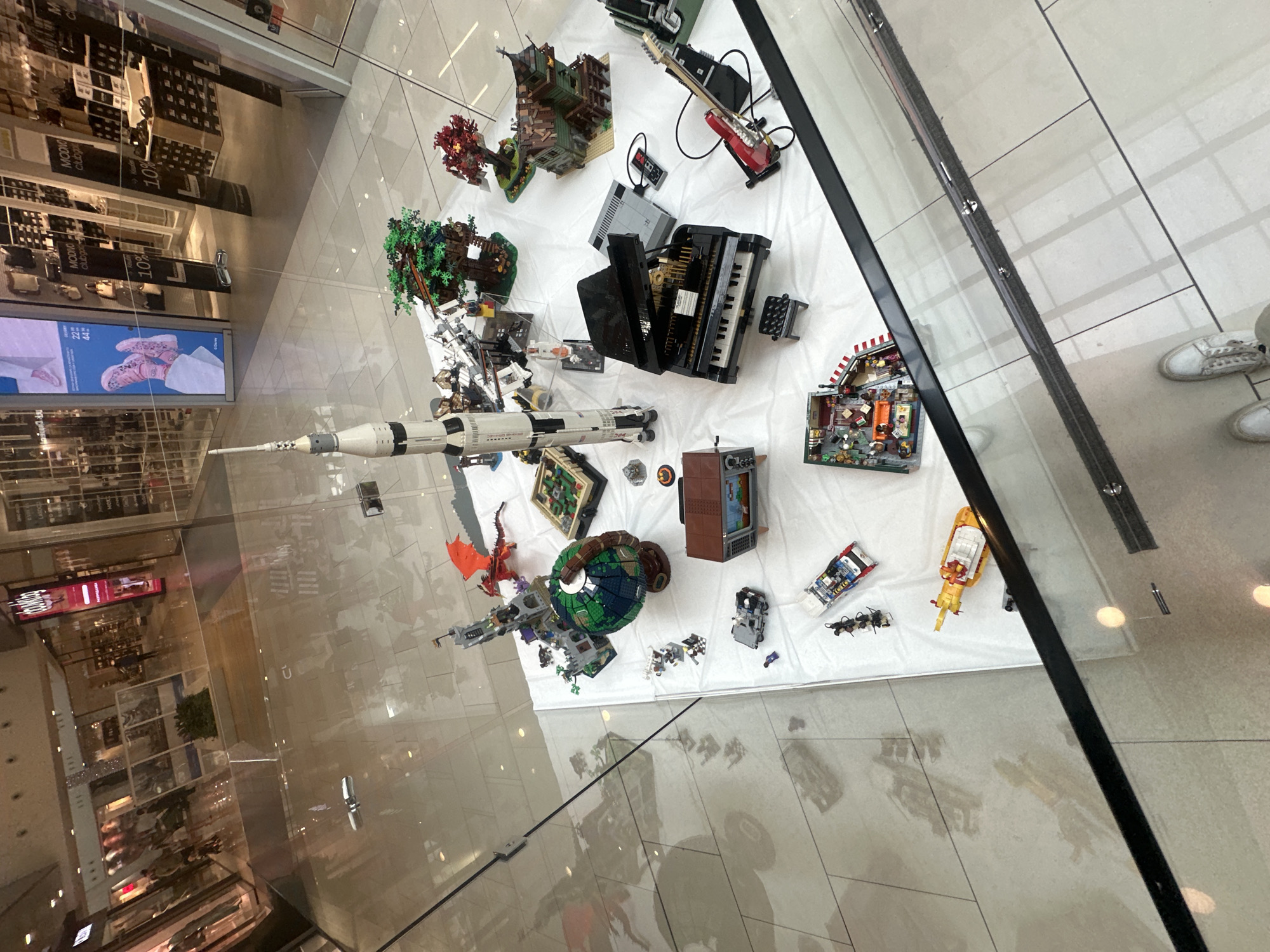 The image shows a display of Lego models on a white surface within a shopping mall. The models include a Saturn V rocket, a castle with a dragon, a globe, a treehouse, a haunted house, a piano, a guitar, a vintage television, a car, a tank, and a building interior. The display is set up on a white cloth table in what appears to be a public area of a shopping mall, possibly in Sofia, Bulgaria, judging by the mall's aesthetic and potential signage not fully visible. The floor is tiled and reflective, showing reflections of the mall's ceiling lights and some distant figures. The overall scene is static, showcasing the intricate Lego creations.