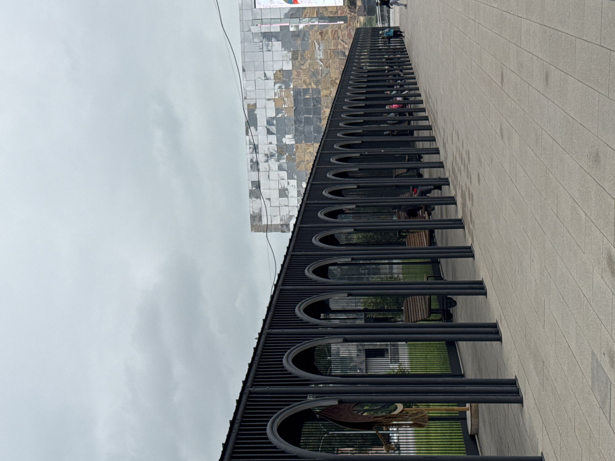 The image shows an outdoor covered walkway with a series of arches supported by dark vertical posts. Benches are visible within the arched sections. Beyond the walkway, a modern building with a reflective, mosaic-like facade stands. The sky is overcast. Several people are present, some seated on the benches and others walking along the paved pathway. The scene is set in the city of Nizhny Novgorod, Russia, specifically on the pedestrian area of the Nizhny Novgorod Stadium.