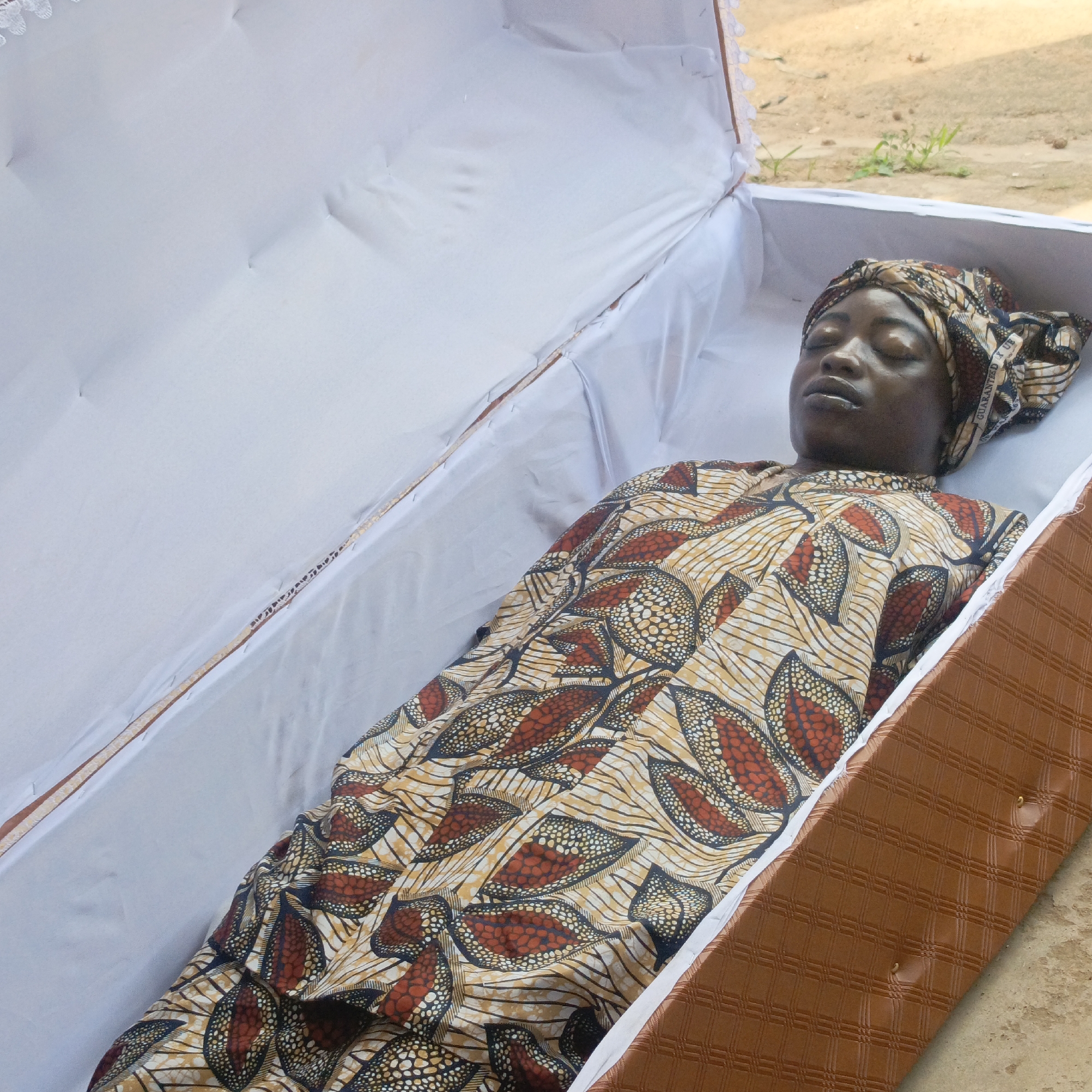 The image shows a deceased dark-skinned woman lying in a casket. She is wearing a patterned dress and headscarf. The casket is lined with white fabric and the exterior appears to be brown. The location is a city named Unknown, Unknown. The environment appears to be outdoors, with a blurred ground surface visible. There are no actions or interactions observed within the image besides the woman lying still in the casket.