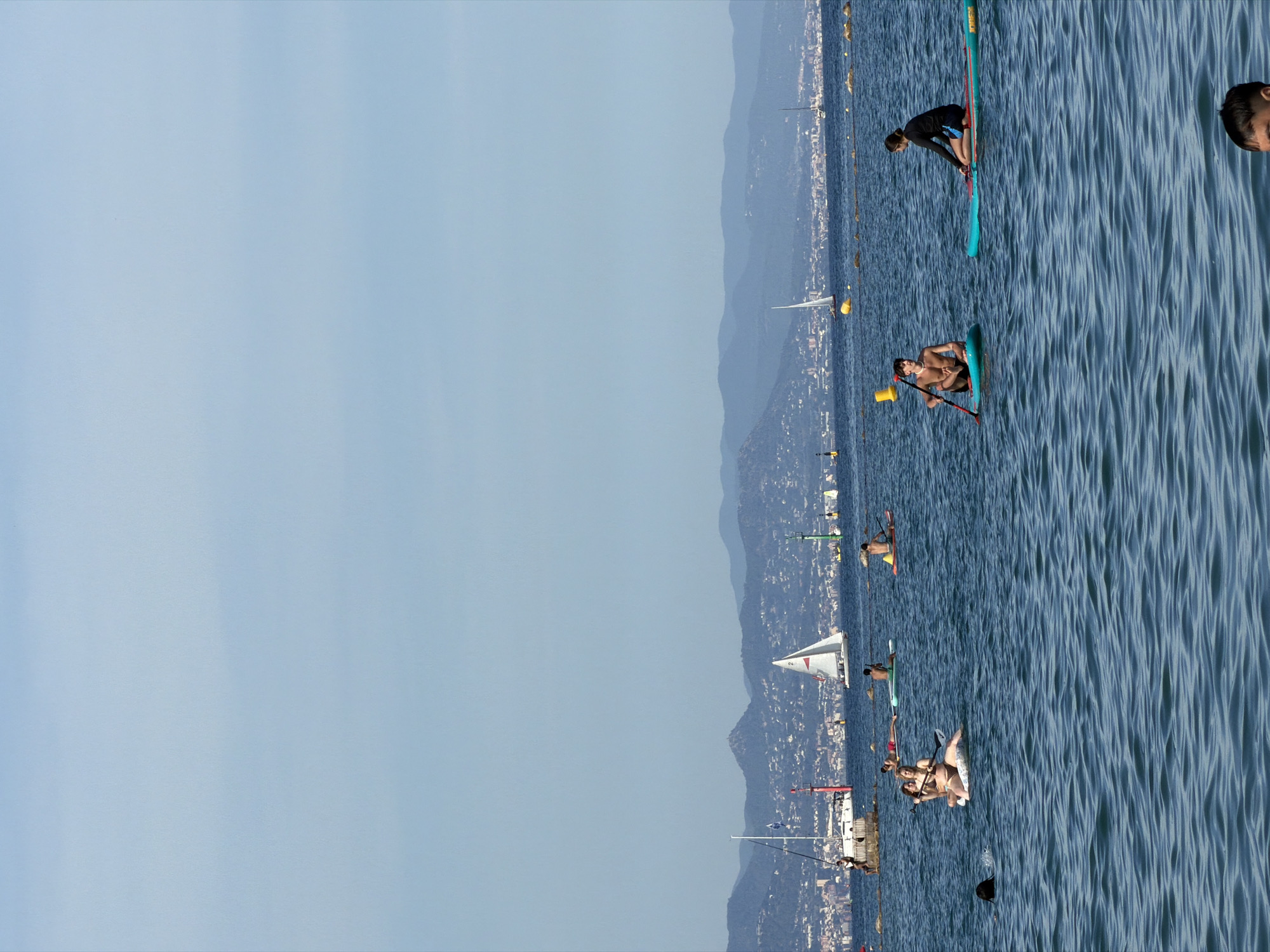 The image depicts a group of people engaged in water activities on a sunny day. The scene is set in the Mediterranean Sea, with Barcelona, Spain, visible in the background. The city skyline, characterized by its numerous buildings and hills, stretches across the horizon, under a clear blue sky. Several sailboats are dotted across the water, some in motion, adding to the nautical atmosphere.
In the foreground, several individuals are on paddleboards. One woman is seated on a paddleboard, holding a paddle, while another person, partially submerged, swims nearby. To the right, two men are also on paddleboards. One man is seated on his board, holding a paddle, and appears to be looking out at the water. Another man, wearing a black long-sleeved shirt and blue shorts, is kneeling on his paddleboard, facing away from the camera. A few other individuals are scattered on paddleboards further out in the water.
The water is calm and appears to be a deep blue, with gentle ripples. The overall impression is one of a leisurely day at the beach, with people enjoying the water and the scenic coastal views of Barcelona.