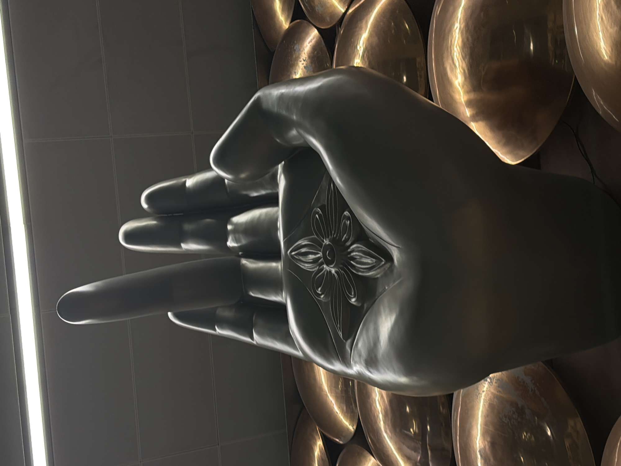 This is a low-angle, close-up photograph of a large, dark-colored sculpture of a hand, likely made of metal, with its fingers extended upwards. The palm of the hand features an embossed floral or lotus-like design. The sculpture is set against a backdrop of numerous dome-shaped, metallic objects, appearing to be golden or bronze, arranged in a mosaic pattern. The lighting suggests an indoor environment, possibly an airport or a public building, with a fluorescent light fixture visible at the top of the frame. The overall impression is one of grandeur and artistry. This sculpture is located in the Indira Gandhi International Airport (DEL) in Delhi, India, specifically in Terminal 3.
