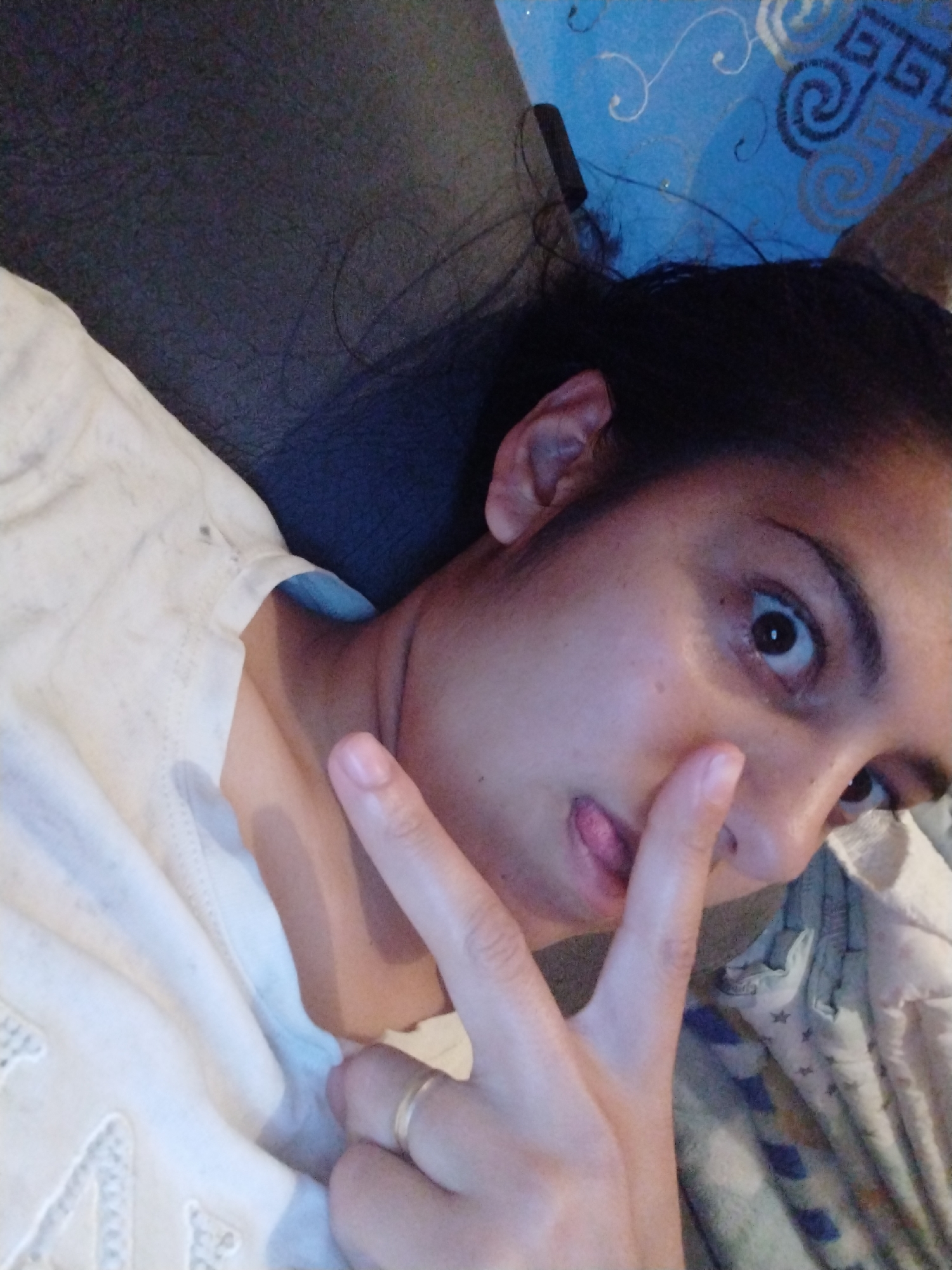 A person is lying on a surface, possibly a couch or bed, with their head tilted towards the camera. They are making a peace sign with their fingers, and their tongue is slightly visible. The person has dark hair and is wearing a white top. The background consists of a blue wall with some patterns and a dark surface, potentially a couch or headboard. The location is Unknown, Unknown.