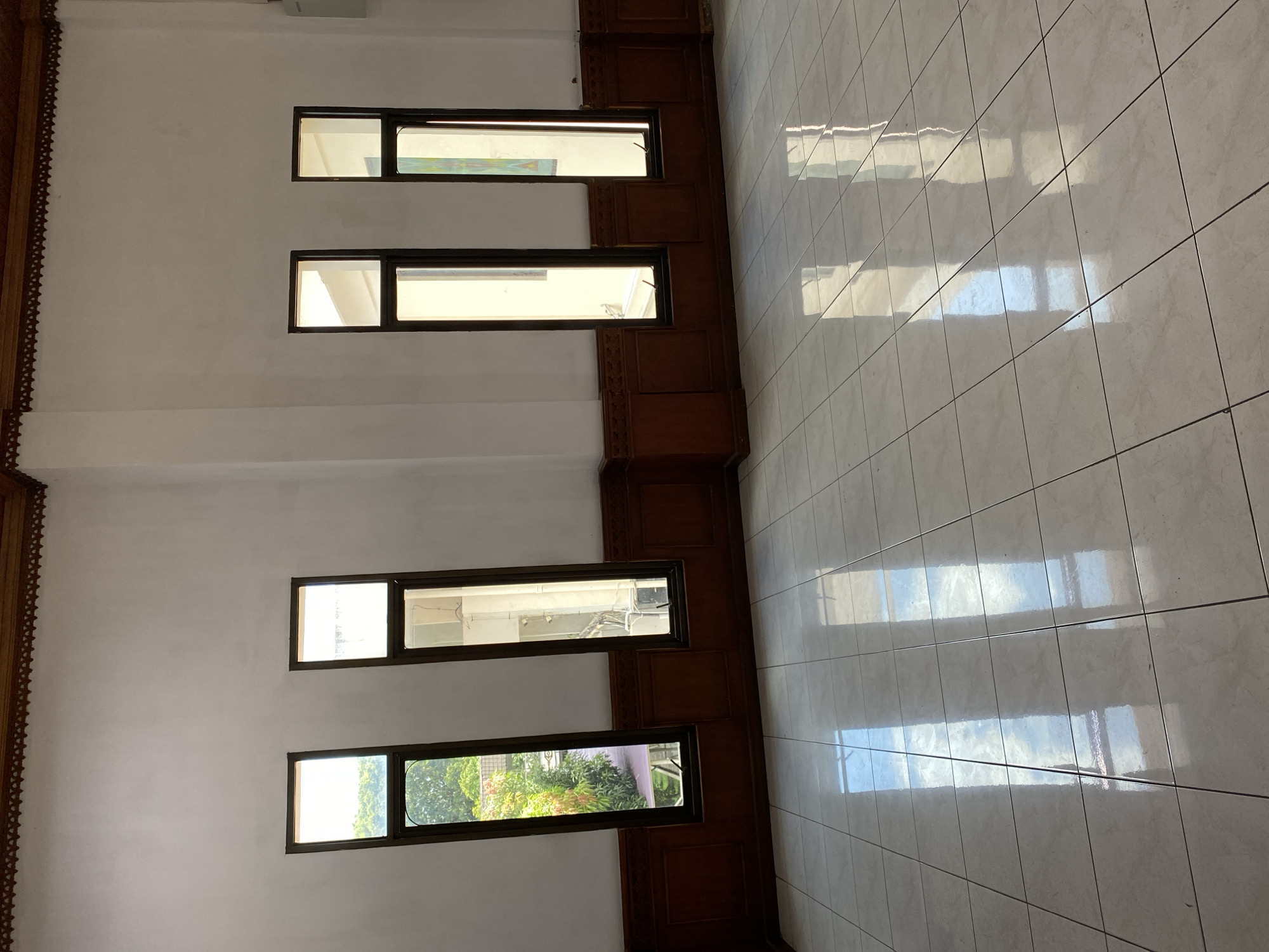 The image depicts an interior room with white walls, a tiled floor, and wooden trim. Four tall, narrow windows line the back wall. The two leftmost windows offer a view of lush green trees and foliage outside, suggesting they overlook a garden or courtyard. The two rightmost windows appear to look into another interior space, with one showing a white wall and the other a colorful painting or artwork. Sunlight streams through the windows, casting reflections on the polished tiled floor. The room appears empty, with no people or furniture visible. The architectural style, particularly the wooden trim along the ceiling and wall, might suggest a traditional Indonesian aesthetic. Considering the mention of Banda Aceh, Indonesia, the scene could be within a public building, a mosque, or a traditional house in that region. The environment is clean and well-lit by natural light.