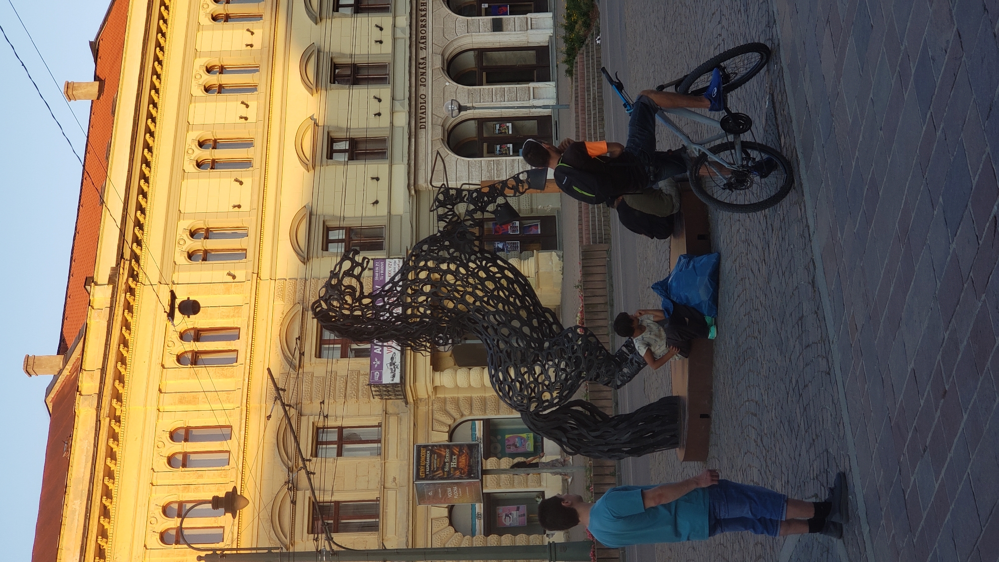 This image captures a street scene in Prešov, Slovakia, likely in front of the Jonáš Záborský Theater. A large, abstract metal sculpture of a horse stands prominently in the foreground, its intricate, woven design catching the light. To the left, a young man in a blue t-shirt and shorts stands facing away from the camera, looking towards the sculpture. Near the base of the sculpture, a child sits on a low platform, dressed in a patterned shirt and dark pants, with a blue tarp bag beside them.
In the background, the facade of a grand, yellow building with classical architectural elements and a red-tiled roof is visible. This building appears to be the theater, with arched doorways and windows. Several posters are displayed on the building's exterior. To the right of the sculpture, a man with a backpack, wearing a black t-shirt with orange accents and dark shorts, sits on a bicycle. His legs are positioned as if he is about to pedal, with one foot on the pedal and the other on the ground. The ground is paved with cobblestones. The lighting suggests it is late afternoon or early evening, with a warm glow illuminating the scene.