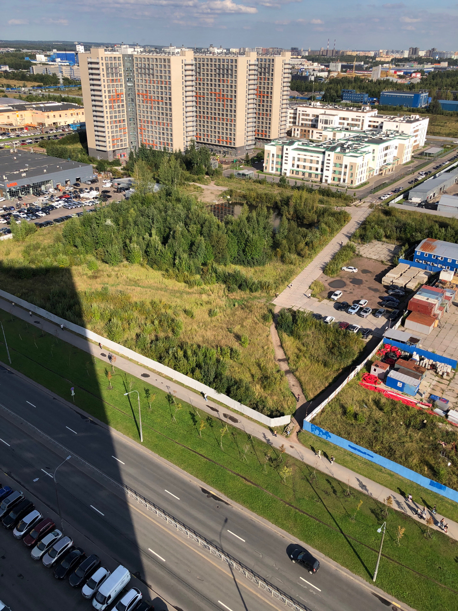 The aerial image shows a large urban development in Lavriki, Russia, featuring high-rise residential buildings and commercial structures. In the foreground, a multi-lane road runs parallel to a grassy embankment and a pedestrian walkway. A line of parked cars is visible along the side of the road. A shadow from a tall building stretches across the road and walkway. Further back, a large, undeveloped grassy area with scattered trees and a small pond is situated between the road and more developed sections of the city. Commercial buildings, including what appears to be a car dealership with many vehicles parked in its lot, are visible on the left. On the right side of the image, there are industrial or storage facilities with shipping containers and construction materials. Several people are observed walking on the pedestrian path. The sky is partly cloudy, indicating daylight.