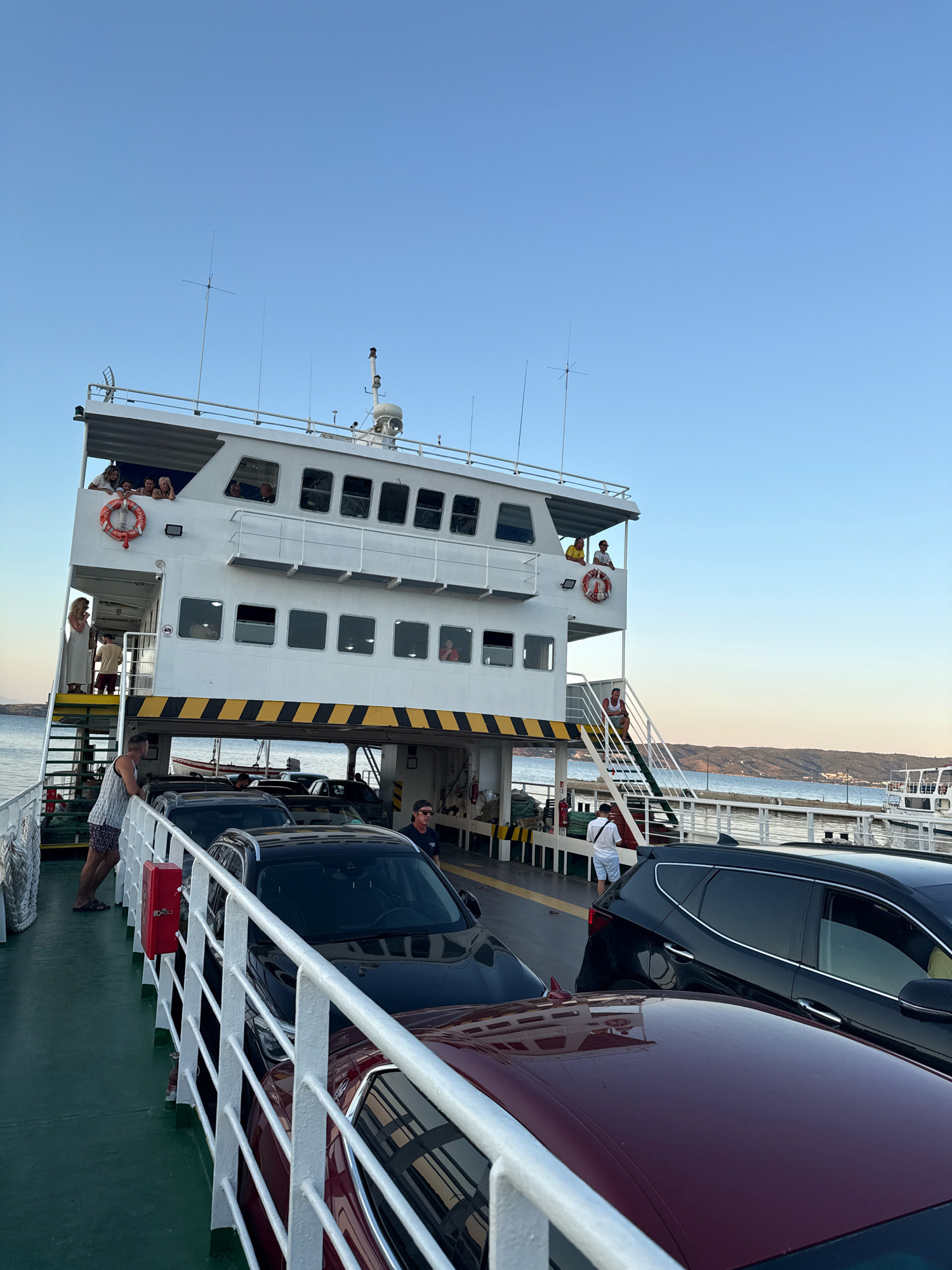 On a ferry in Ammouliani, Greece, cars are parked in rows on the main deck. The ferry has multiple levels, with passengers visible on the upper decks looking out. The sky is clear and blue, with a hint of sunset colors on the horizon. The water is calm, and the ferry is docked or moving slowly near a shore with hills in the distance. People are standing or sitting on the ferry, some observing the surroundings, while others are near their vehicles. The ferry appears to be white with yellow and black hazard stripes. There is a red fire extinguisher mounted on a railing.