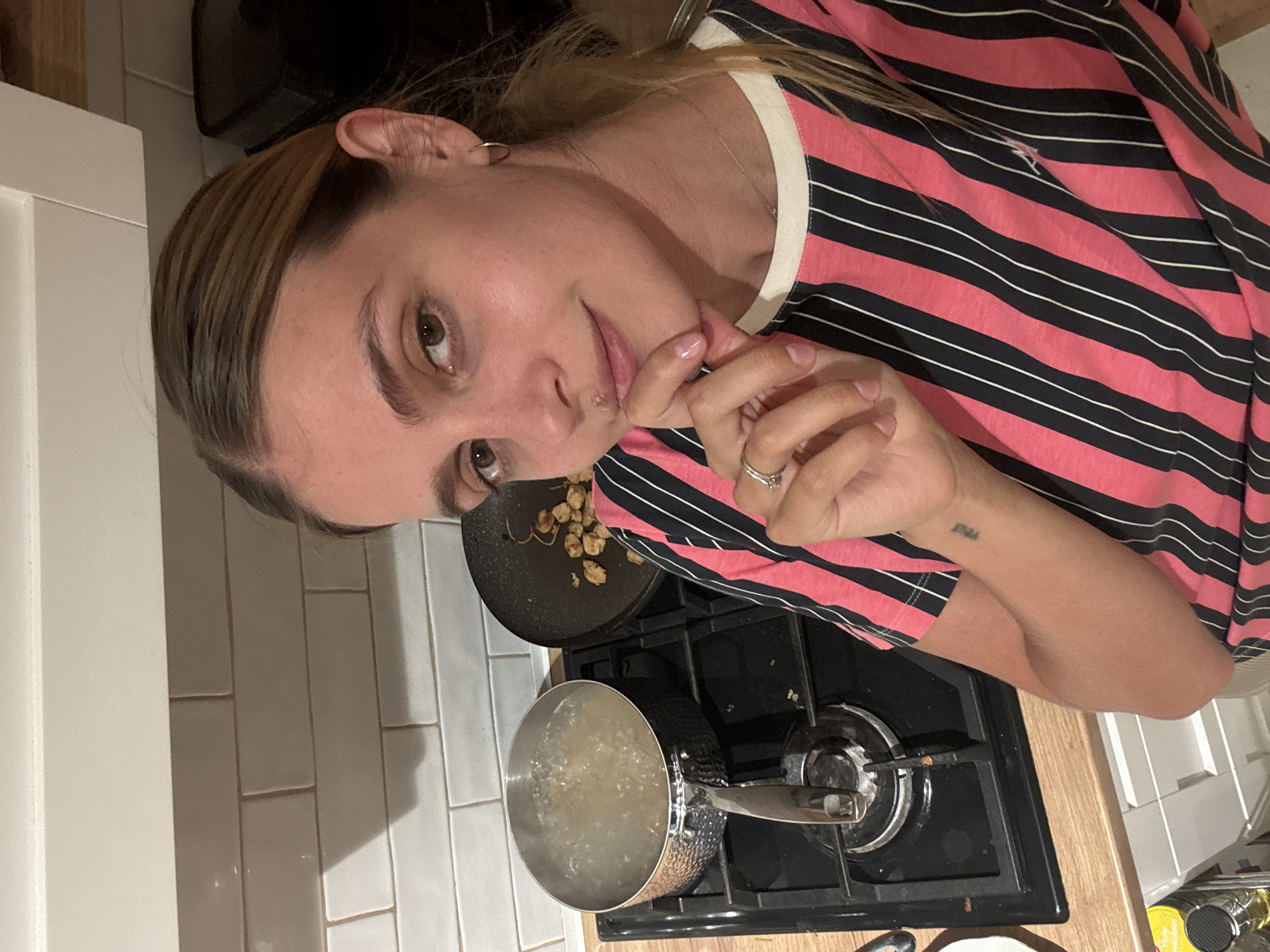 A woman with light brown hair and prominent highlights is captured in a close-up, rotated portrait. She wears a pink t-shirt with thin black and white horizontal stripes and a cream-colored collar. A delicate silver chain is visible around her neck, and a small silver hoop earring adorns her left ear. She has a silver ring on her left ring finger and light pink nail polish. Her left hand is raised, with a finger resting near her mouth, and a small lesion is visible on her upper lip. A small tattoo, possibly reading "STILL", is visible on her left wrist. Her expression is calm, and she gazes directly at the camera.
The setting is a kitchen, identifiable by a gas stove with two burners. On the front burner, a silver pot with a textured exterior is heating, containing liquid that appears to be boiling. To the right, a black frying pan is partially visible, holding what looks like cooked, crumbled food. The background features a white tiled backsplash and a wooden countertop. The lighting suggests an indoor setting, likely evening or artificial light. The woman appears to be pausing from cooking to pose for the photo.
