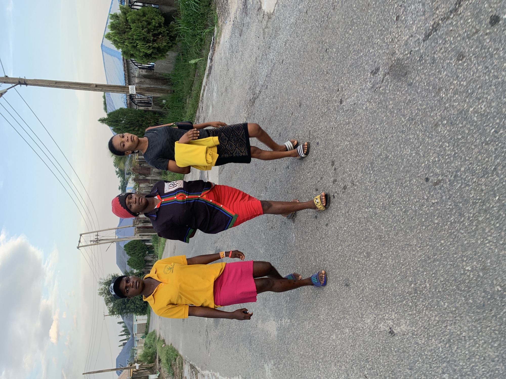 Three individuals are walking along a paved road.
The person at the bottom is wearing a yellow polo shirt, a pink skirt, blue sandals, and a baseball cap.
The middle person is wearing a black long-sleeved shirt, red shorts, and a red beanie hat.
The person at the top is wearing a black t-shirt, a black skirt, white and black slides, and is carrying a yellow garment.
There are buildings and vegetation in the background. Utility poles are visible on the left of the frame.
The location is Abuja, Nigeria.