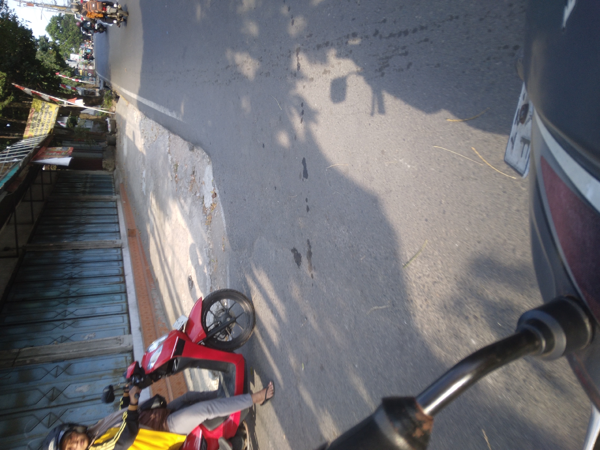 The image shows a street scene in Sokaraja, Indonesia, with a red scooter in the foreground, being ridden by a person wearing a yellow jacket, grey pants, and a helmet. The scooter is moving on a paved road, with a sidewalk and buildings on the left side of the frame. The buildings appear to be shops, with closed metal shutters. Trees are visible on the right side of the road, casting shadows on the pavement. Other scooters and motorbikes are visible in the background, indicating traffic. The time of day appears to be late afternoon, as indicated by the long shadows.