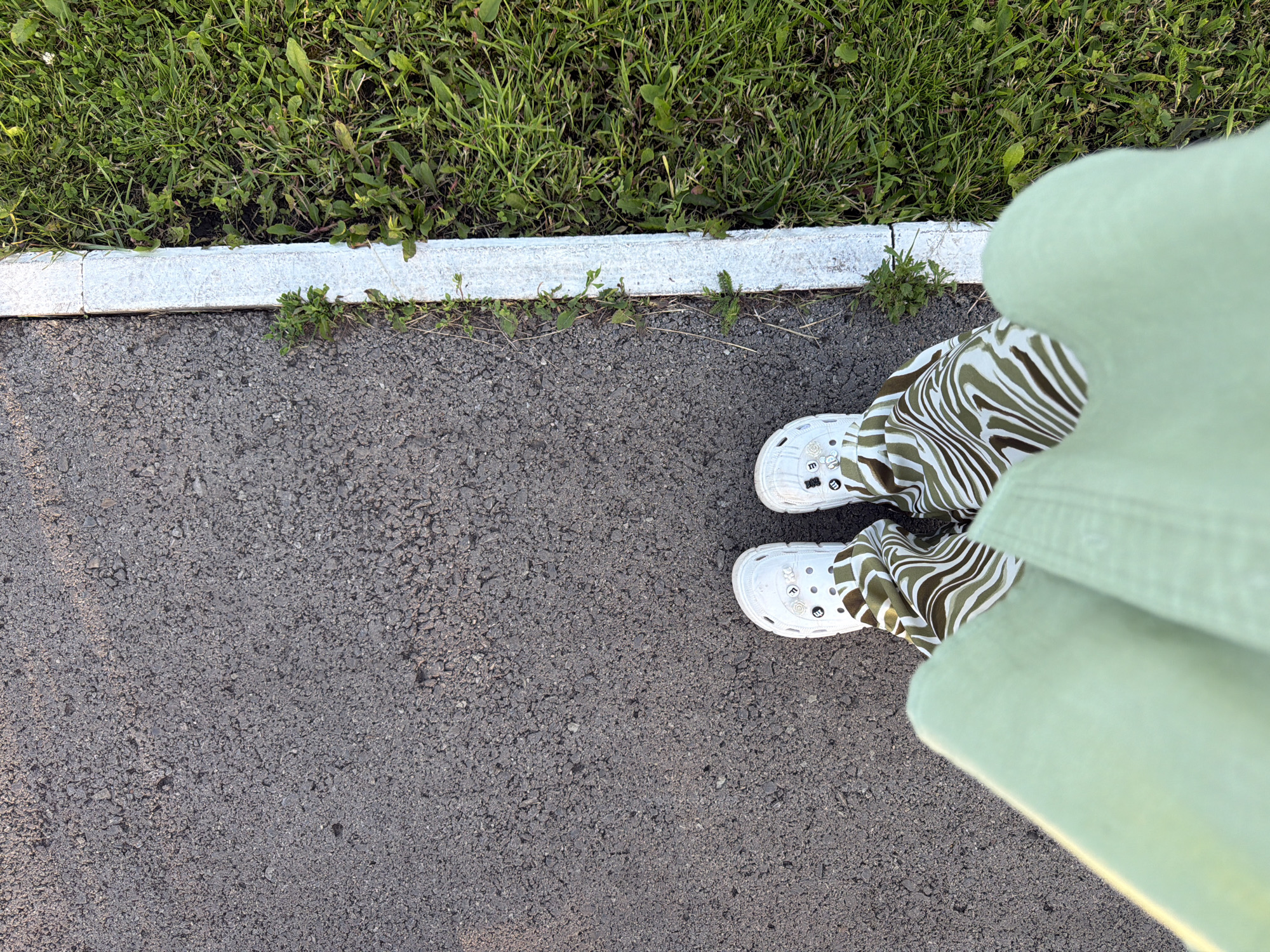 In Шмидтово, Russia, a person stands on an asphalt surface next to a white painted curb, with green grass bordering the curb. The person is wearing white Crocs and pants with a green and brown wavy pattern. The image is taken from a top-down perspective, focusing on the lower body and surroundings.