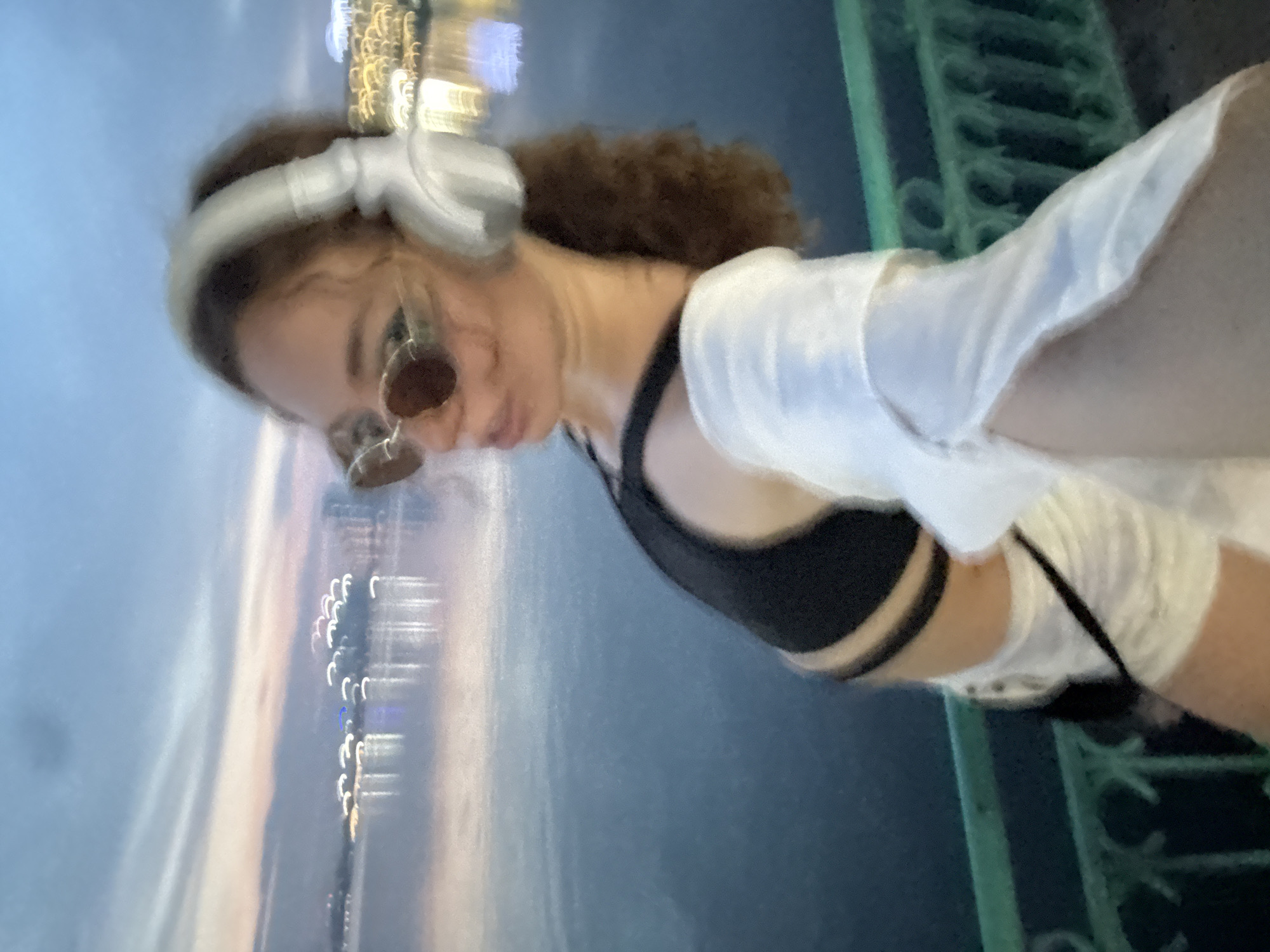 The image shows a young woman with curly brown hair wearing headphones and sunglasses. She is wearing a black sports bra and a white shirt draped over her shoulders. She appears to be outdoors at night, with blurred city lights in the background. The lights suggest a waterfront location with buildings or structures illuminated across the water, possibly reflecting on the surface. A green railing is visible on the right side of the frame, indicating she might be on a balcony, bridge, or promenade. The overall mood is relaxed and contemplative, with the city lights and headphones suggesting an urban, possibly musical, experience. The scene is set in Hanoi, Vietnam, and the time appears to be evening or night. There is no visible text in the image.