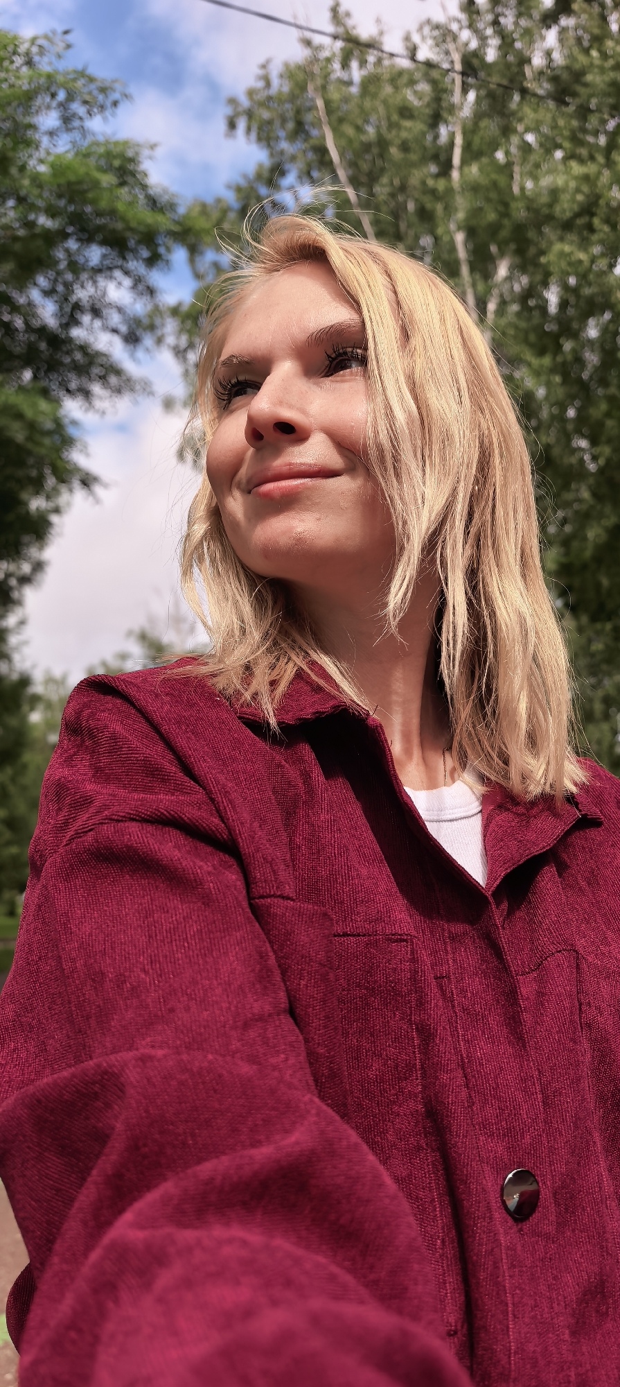 A young blonde woman is captured outdoors in Sterlitamak, Russia, viewed from a low angle. She has a fair complexion with visible freckles on her nose and cheeks, and is looking upward and to the right of the frame with a gentle, content smile. Her medium-length blonde hair is styled casually, appearing slightly wavy with some flyaway strands. She wears an oversized, deep crimson corduroy shirt-jacket, characterized by prominent vertical ridges in the fabric, layered over a white crew-neck t-shirt. A dark, circular button is visible on the jacket's placket in the lower right portion of the image. The background features a clear blue sky with scattered white clouds and dense green foliage from trees, suggesting a natural environment. A thin, dark line, possibly a utility cable, is visible traversing horizontally across the upper background between the trees.