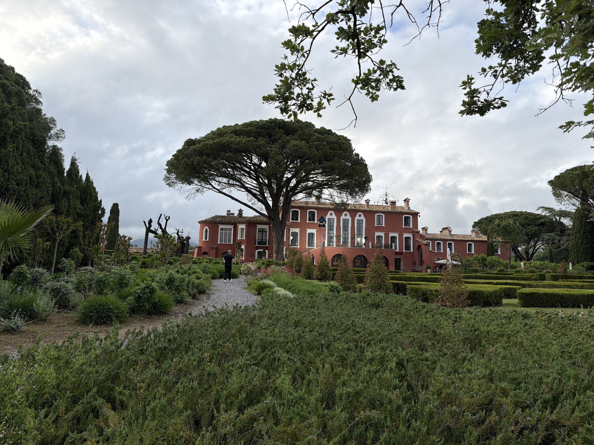 The image shows a large, red building surrounded by manicured gardens. The location is in Cogolin, France. In the foreground is a dense patch of green shrubs. A gravel path leads towards the building, where a man in dark clothing is walking. The building is two stories tall with white-framed windows and a red tile roof. A large tree stands in front of the building, partially obscuring the facade. In the background are various trees and hedges, some forming a maze-like pattern. The sky is overcast and cloudy.