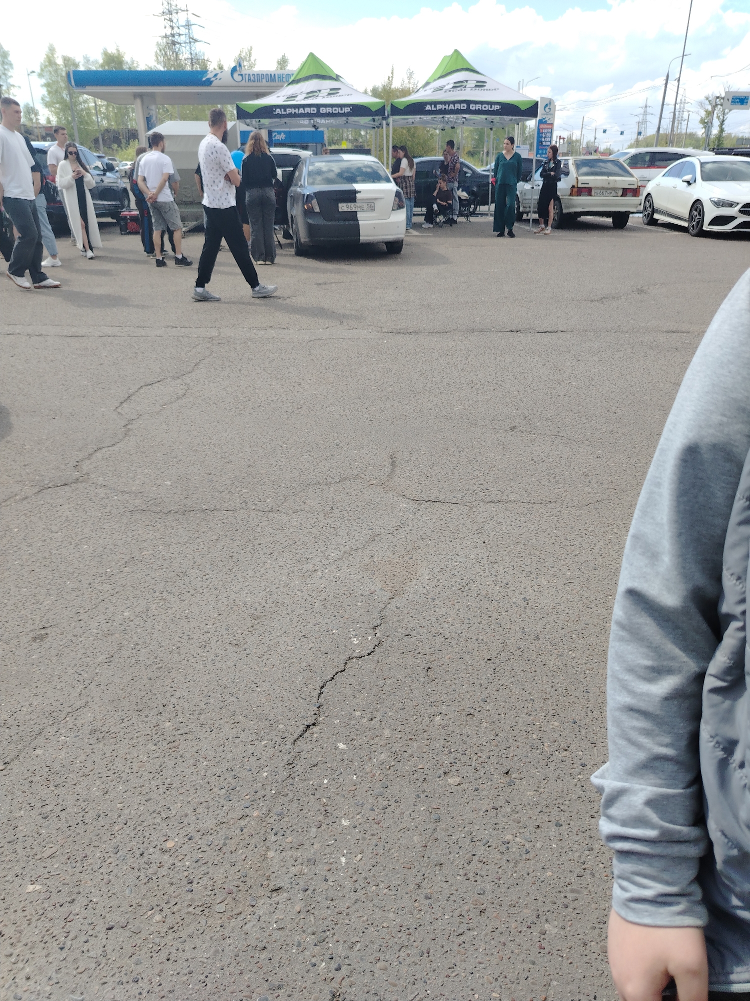 The image depicts a scene in what appears to be a parking lot in Шебунино, Russia. Several individuals are present, standing and walking around a group of parked cars. In the background, there is a Gazprom Neft gas station and tents labeled "Alhard Group." The atmosphere suggests a casual gathering or event. The foreground shows cracked asphalt and a person's arm wearing a gray sweatshirt.