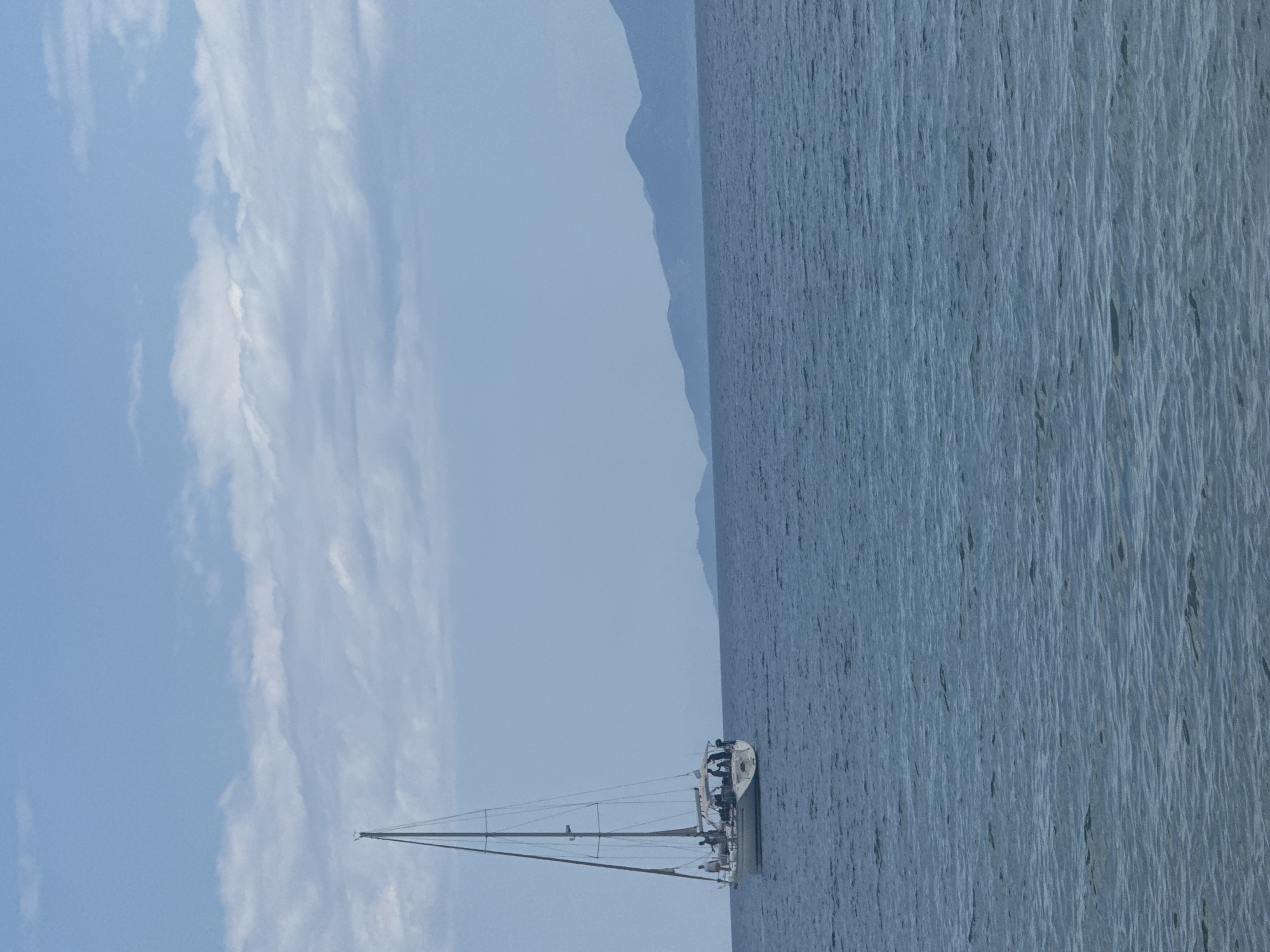 The image depicts a sailboat sailing in the Aegean Sea near Kavala, Greece. The sea is a light blue-gray, and mountains are visible in the background. The sky is mostly clear with some light white clouds. A few people appear to be on the sailboat. The image is oriented with the horizon running vertically.