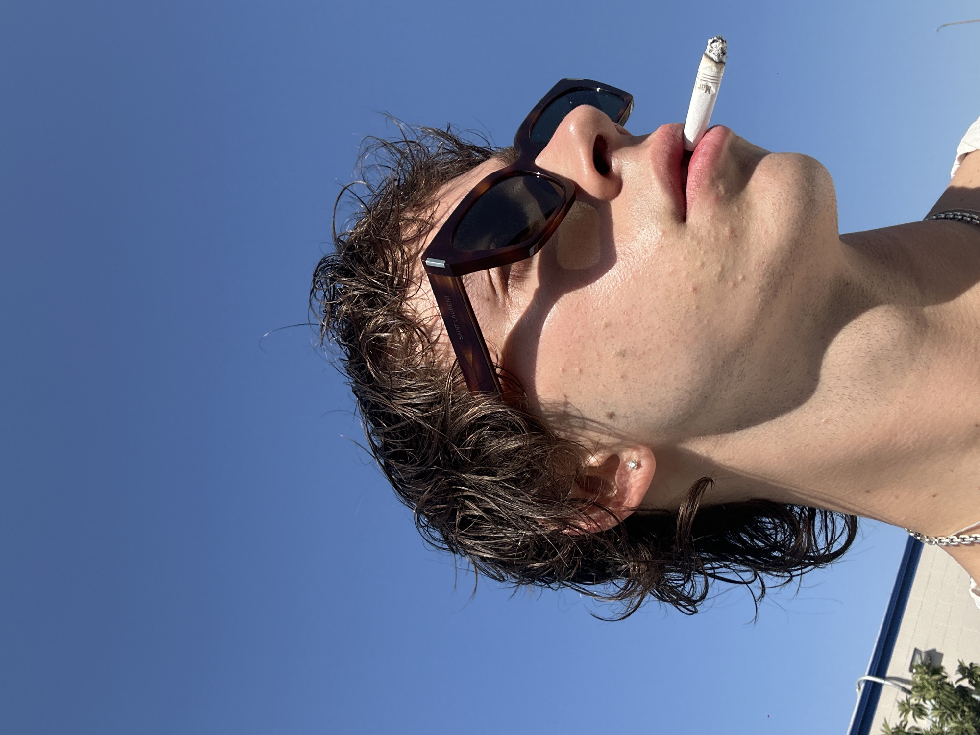 A medium shot depicts a person from a low angle, looking up at the clear blue sky. The person has dark, wet hair, sunglasses, and is holding a lit cigarette in their mouth. A building is partially visible on the right side of the image, along with some vegetation. The sunglasses are Saint Laurent sunglasses. The cigarette has a brand name, "Marlboro", on the side of it. The location is assumed to be somewhere in Glendale, United States of America, given the prompt.