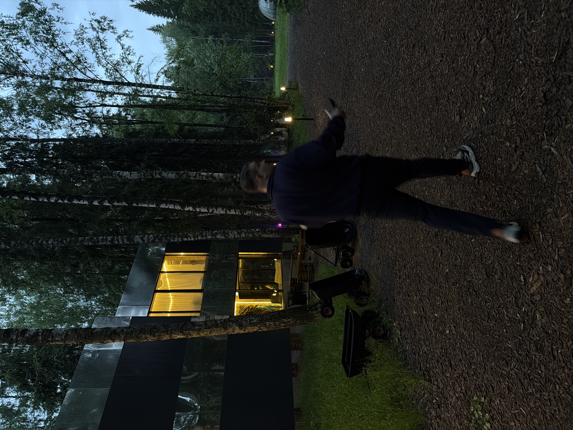 The image depicts an outdoor scene at night in what appears to be a park or wooded area. There is a blurred person walking on a pathway paved with dark material, possibly mulch or wood chips. To the left of the pathway is a modern-looking building with a dark facade, possibly made of reflective panels, and windows illuminated from within with warm light. Several small trailers or carts are also visible. In the background, there are trees and faint lighting in the distance. The image could be from Bakhtiyary, Russia, given the prompt information.