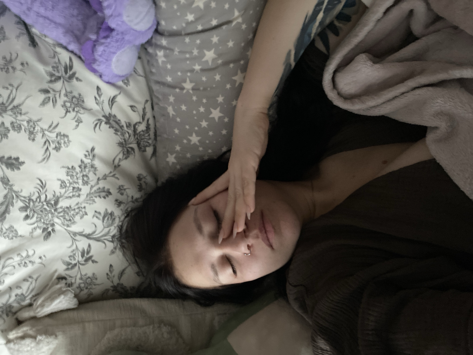 A person lies in a bed covered with bedding, including a floral-patterned sheet, a star-patterned pillow, and a gray blanket. The person's left hand is resting on their face, covering their forehead. The person has dark hair and a nose ring. A tattoo is visible on the person's left arm. A purple plush object is visible on the bed. The location of the image is unknown.