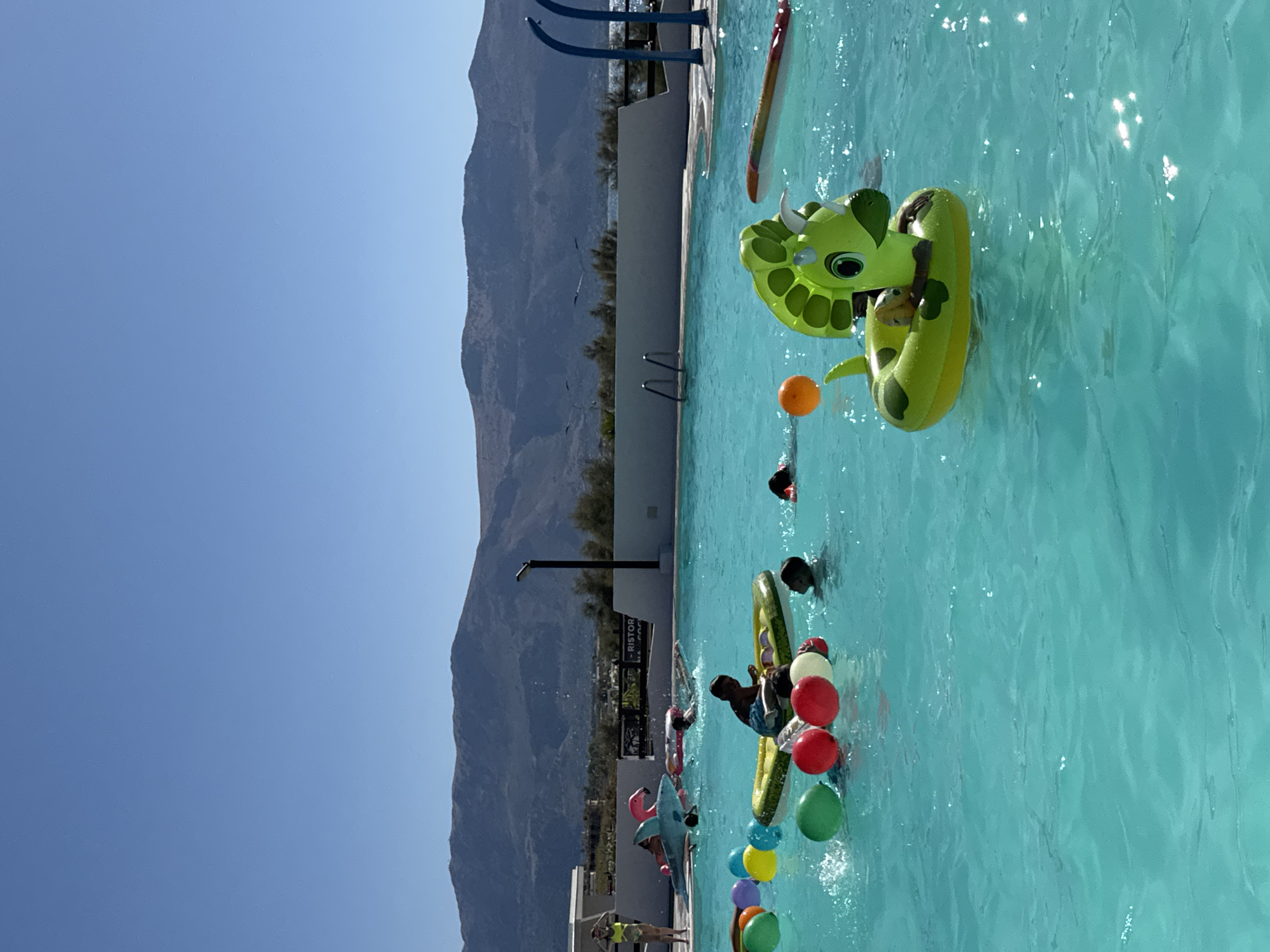 The image depicts a sunny day at an outdoor swimming pool, likely a resort or hotel, located near the city of Capaci, Italy. In the foreground, several children are enjoying the water. Some are swimming, while others are interacting with various inflatable toys, including a green dinosaur-shaped float with a child inside, a long inflatable float with a colorful pattern, and an inflatable boat carrying at least one child. A cluster of colorful balloons, including red, green, yellow, and orange, floats on the surface of the turquoise water. In the mid-ground, more people are visible, some swimming and others on floats, including a pink flamingo float. The pool area is bordered by a white wall, with buildings and trees visible beyond. In the background, a range of mountains forms the backdrop against a clear blue sky. The overall scene is one of recreation and leisure in a warm, Mediterranean environment.