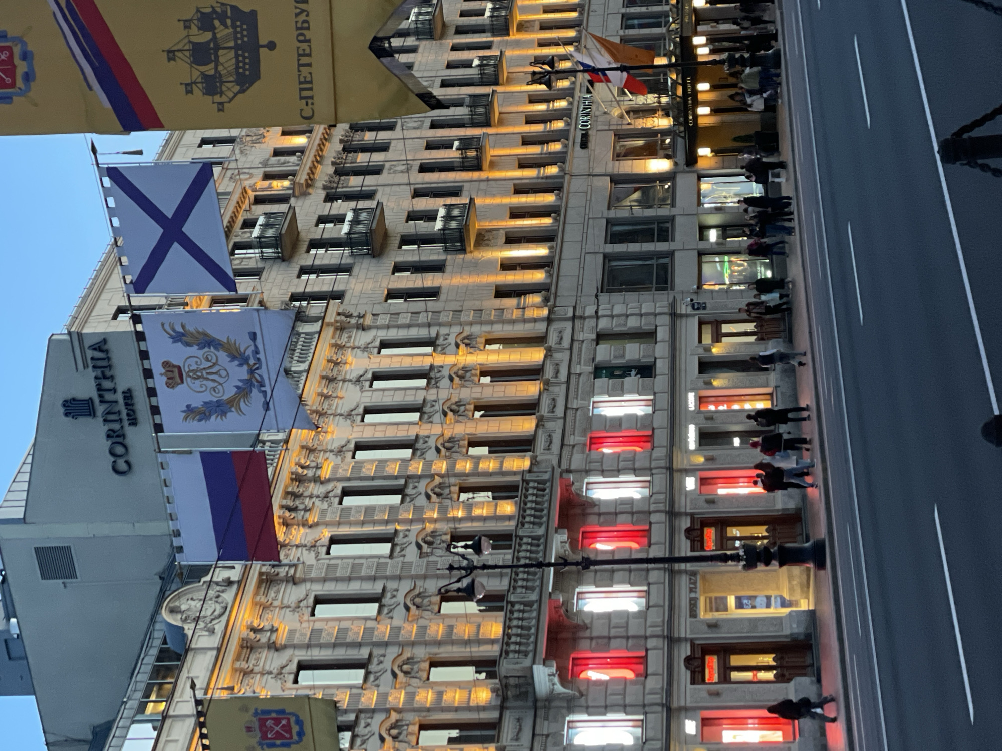 This image, captured at dusk or night in Saint Petersburg, Russia, presents a vibrant street scene centered on a large, ornate multi-story building. The architecture features classical details and numerous windows. A prominent sign identifies the building as the "CORINTHIA HOTEL." Many windows on the upper floors are illuminated with a warm, golden light, while several on the lower floors glow distinctly in alternating red and white hues, adding a dynamic visual element to the facade.
Various flags adorn the building and stretch across the street. These include the national flag of Russia (white, blue, and red horizontal stripes) and the St. Andrew's flag (a white field with a blue saltire), which is the historical ensign of the Russian Navy. Additionally, there are yellow decorative banners; one prominently displays a sailing ship and the text "С-ПЕТЕРБУРГ" (S-Petersburg), while another features a crowned coat of arms.
On the ground level, a wide sidewalk runs alongside the building, populated by numerous pedestrians. People are seen walking and standing, presumably in front of shops or establishments. The street itself is an asphalt road with clear white lane markings, appearing relatively clear of traffic directly in front of the hotel. The artificial lighting and active sidewalk suggest an evening urban environment.