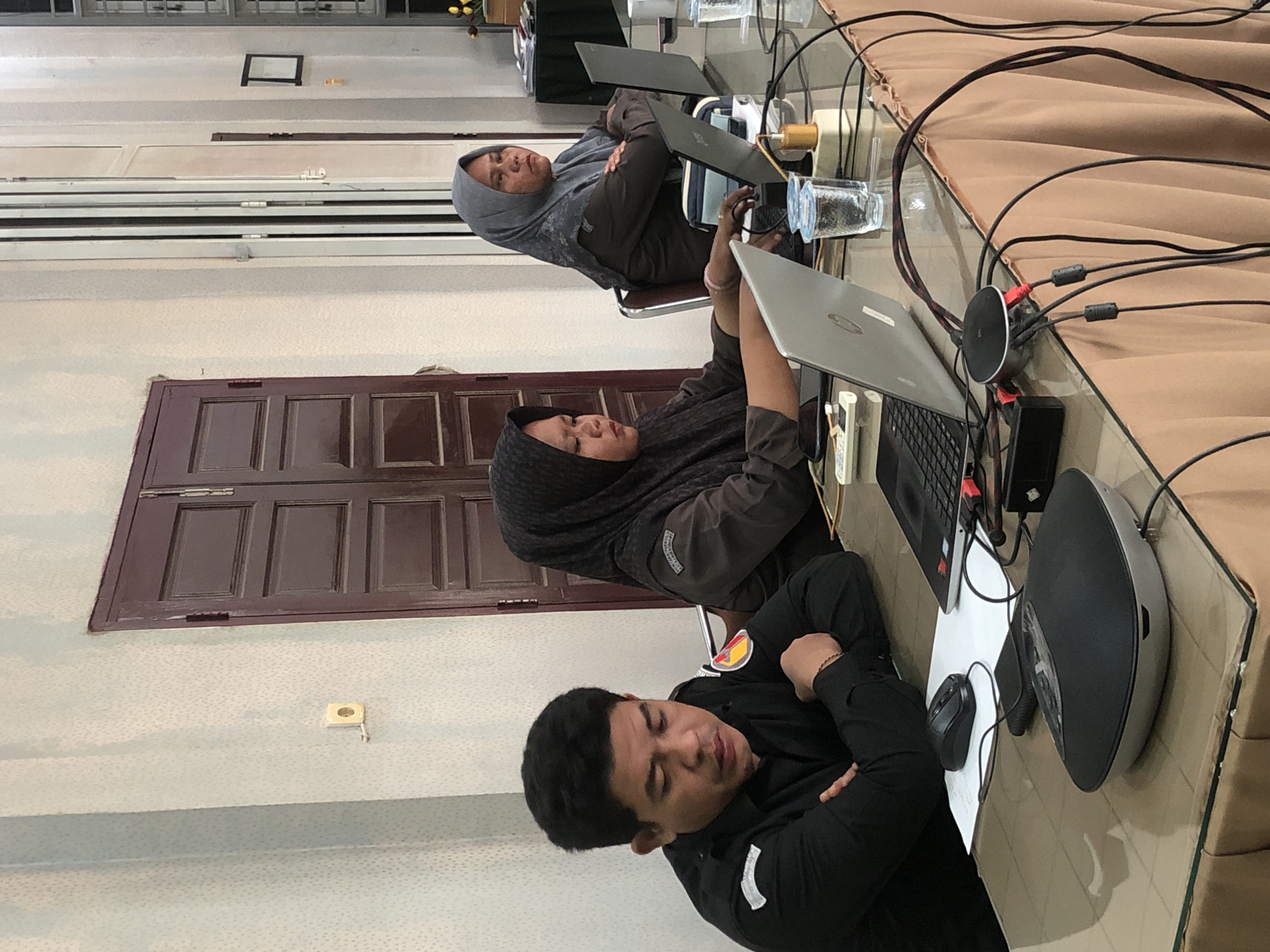 A man wearing a black uniform shirt is seated at a glass-topped table, looking down. To his right, two women in hijabs are also seated at the table. The woman closer to the viewer is looking down at a laptop, while the woman further away is looking forward with her arms crossed. Several laptops, glasses of water, and electronic devices are on the table. The background shows a room with light-colored walls, a dark-colored double door, and a closed-off area with metal folding doors. The image appears to have been taken in Lhokseumawe, Indonesia, possibly in an office or meeting room.
