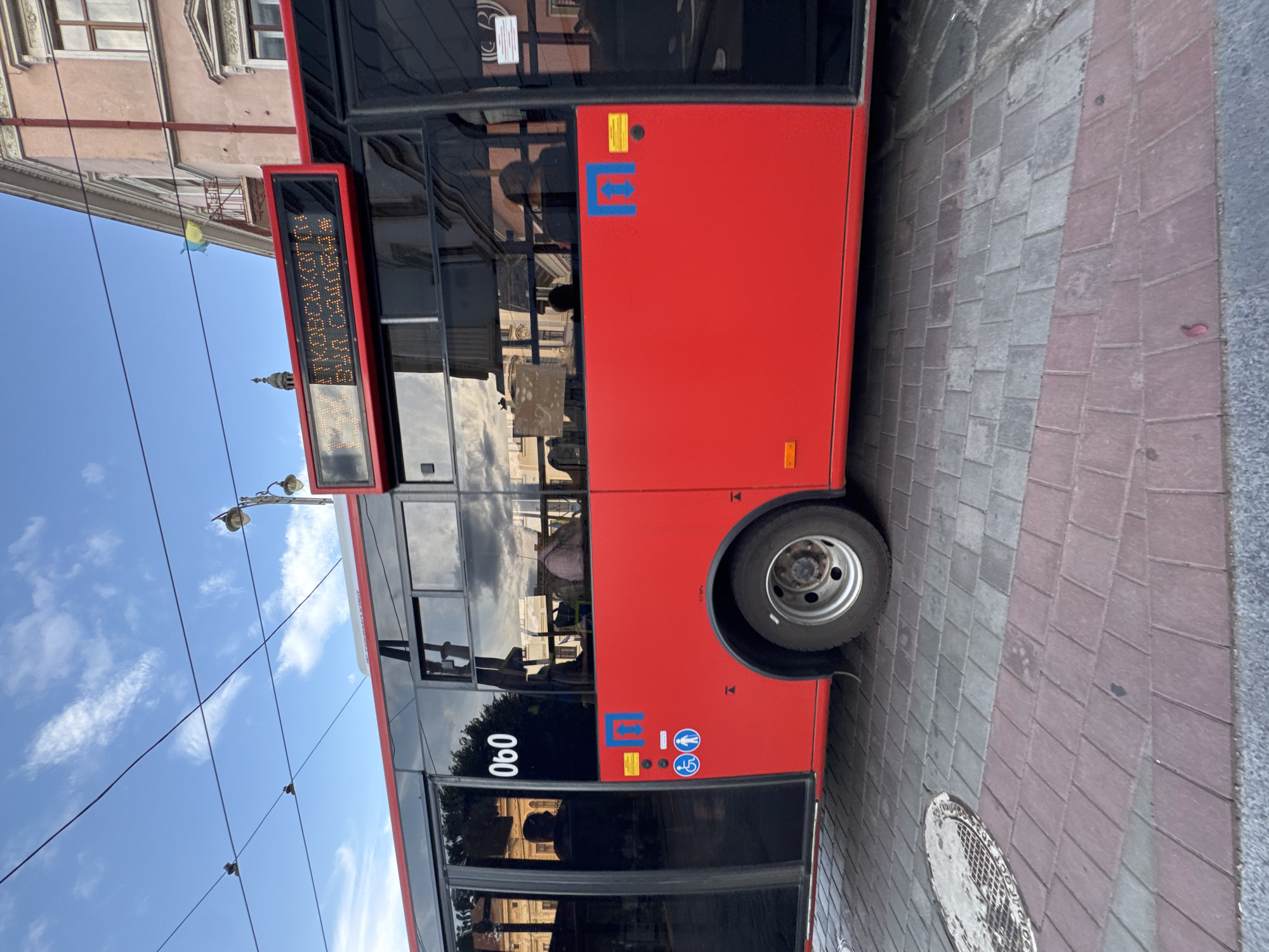 This is a portrait-oriented, sideways shot of a red public bus parked on a street with a cobblestone and brick pavement. The bus has tinted windows and a digital display board at the front showing text in Cyrillic, which appears to read "13:30": "КОРОВСЬКА", "КОВІСЬКИЙ", "МАСАЛТИН". The bus also has various symbols on its side, including a blue arrow pointing left and right and a wheelchair accessibility icon. The number "060" is visible in white on the side of the bus.
In the background, a light-colored building with windows and architectural details is partially visible. A Ukrainian flag, yellow and blue, is hanging from a pole or wire. The sky above is blue with scattered white clouds, suggesting it is daytime. Overhead wires and streetlights are also present. The overall scene suggests a typical urban environment in Chernivtsi, Ukraine, possibly during the day given the clear sky. There are reflections visible in the bus windows, but they are indistinct. The image composition is unusual due to the sideways orientation.