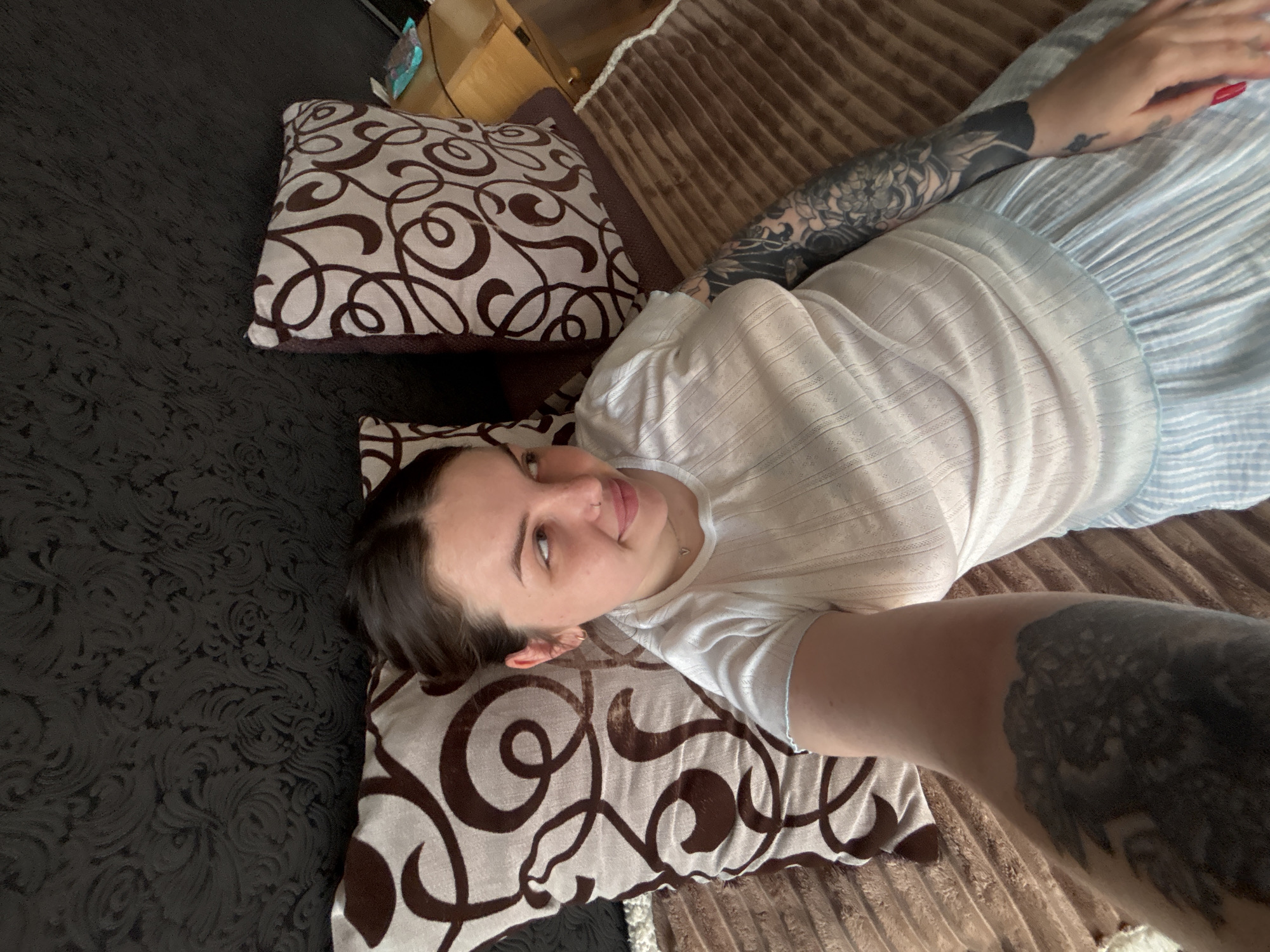 A woman with dark hair lies on a bed with brown patterned pillows. She wears a white, short-sleeved shirt and a light blue and white striped skirt. Tattoos cover her left arm. The bed is covered with a textured brown blanket. To the left, the bed extends onto a dark, patterned surface. The location is unknown but possibly Chornomorskoe, Ukraine.