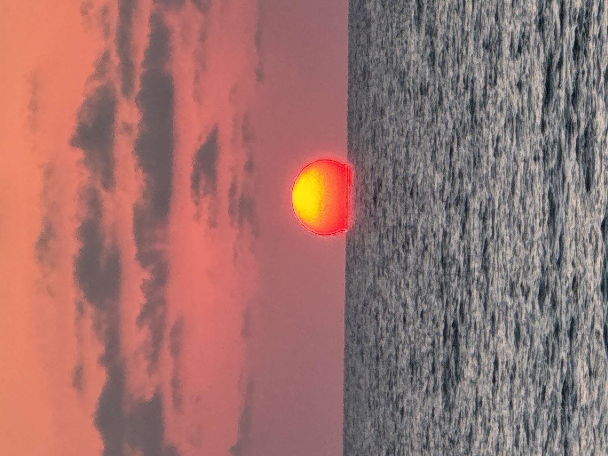 A vibrant sunset over the Mediterranean Sea is captured in this image, likely taken from a beach or promenade in Tel Aviv, Israel. The sun, a brilliant orb of orange and yellow, is partially submerged below the horizon line, casting a warm glow across the sky. The sky itself is a gradient of soft pinks and oranges, with scattered grey clouds adding texture and depth. The sea is calm, with gentle ripples reflecting the colors of the sunset. The overall impression is one of tranquility and natural beauty.