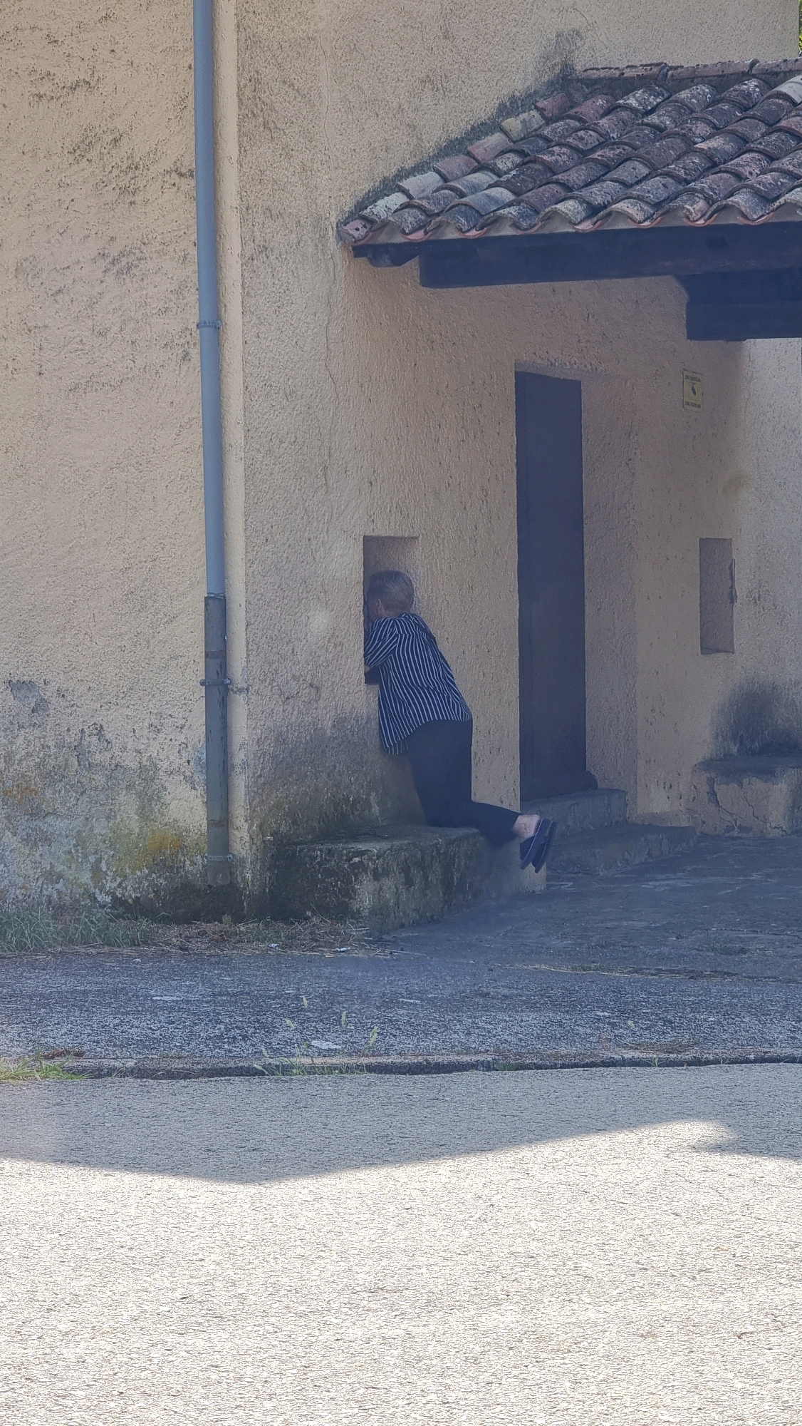 A person is kneeling on a stone ledge and peering through a small, rectangular opening in the stucco wall of a building. The person is wearing a striped long-sleeved shirt and dark pants. The building has a tiled roof and a dark doorway. The ground in front of the building is paved. The scene appears to be in a rural or semi-rural area in La Vall de Bianya, Spain, as indicated by the rustic architecture and natural surroundings.