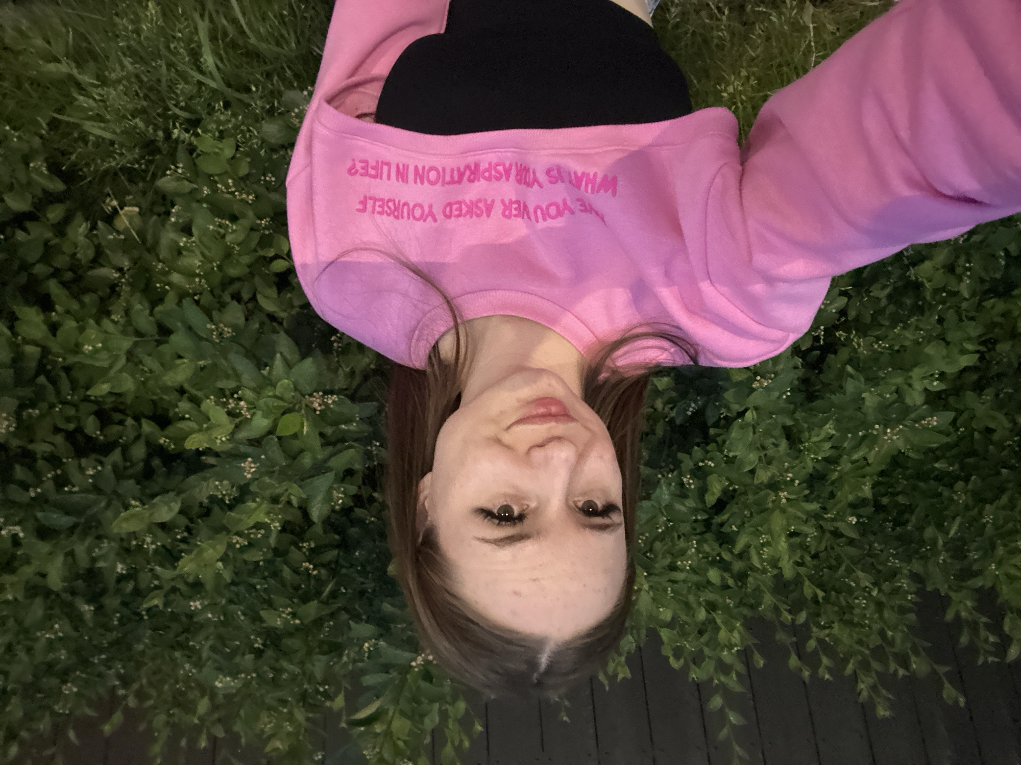 A young woman with brown hair, wearing false eyelashes, is taking a selfie outdoors at dusk. She is wearing a pink cropped sweatshirt over a black top. The sweatshirt has pink lettering that reads, "HAVE YOU EVER ASKED YOURSELF WHAT IS YOUR ASPIRATION IN LIFE?". She is standing in front of a dense green bush and a dark wooden fence.