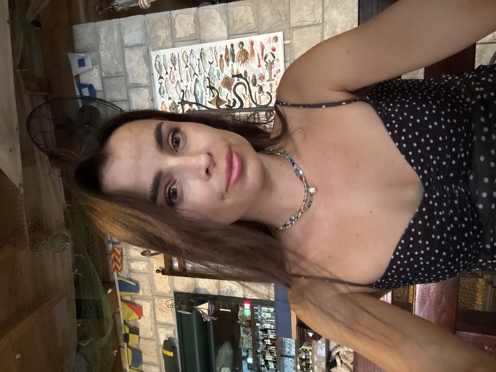 A woman is pictured indoors, appearing to be at a restaurant or bar. She is seated and looking at the camera, with her head slightly tilted. The background includes a brick wall decorated with nautical flags and a poster of various fish species. To the left, shelves with bottles and glassware suggest a bar area. A ceiling fan is visible at the top right of the image. She is wearing a black polka dot dress and a beaded necklace.