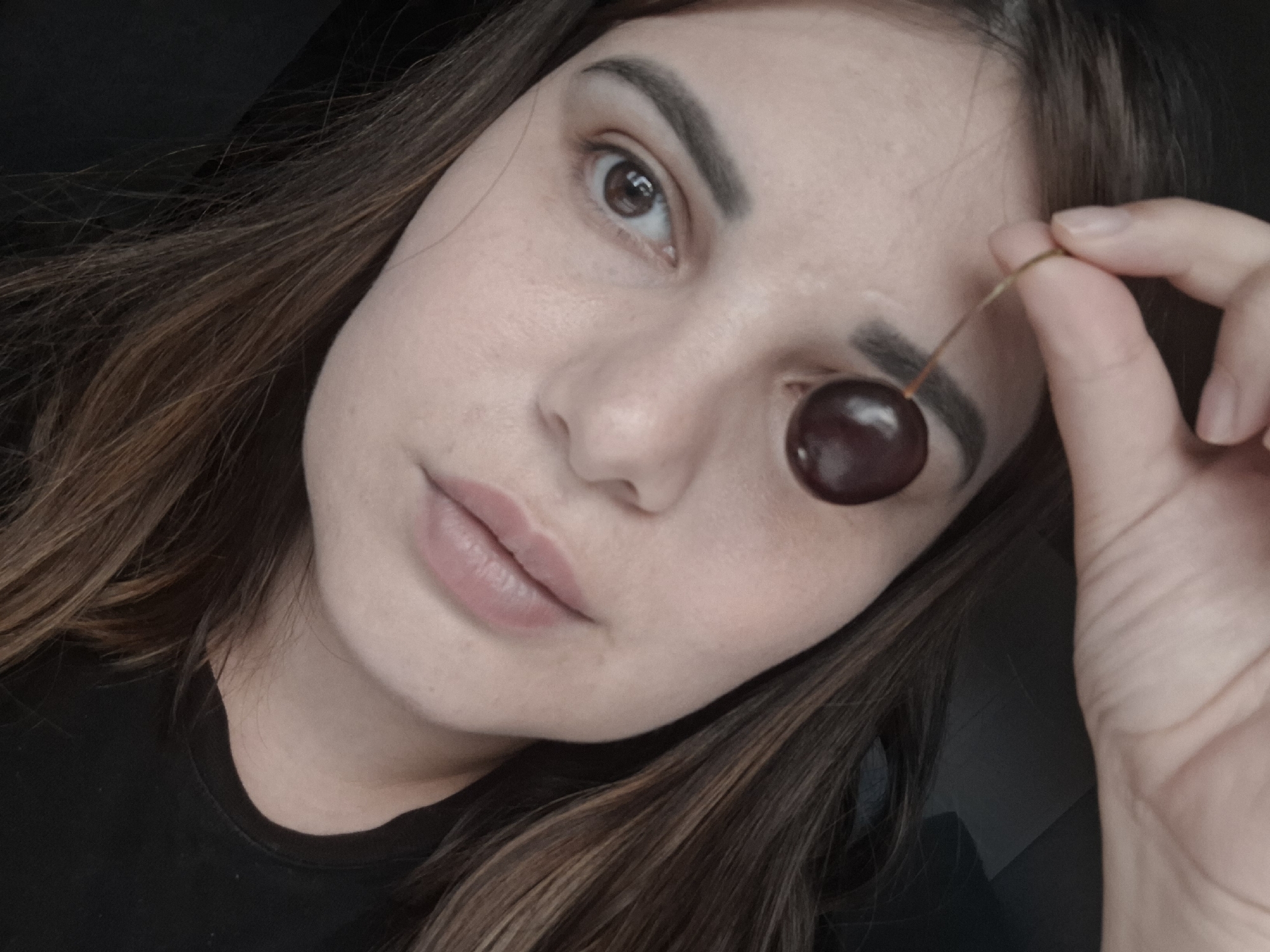 A young woman of East Asian descent is holding a cherry in front of her eye, with the stem extending towards her fingers. The cherry is dark red and slightly oval-shaped, with a subtle sheen. Her skin is fair and smooth, with visible pores and a hint of natural blush. Her eyebrows are dark and well-defined, framing her brown eyes which have a natural, subtle makeup application. Her lips are full and appear to be naturally colored, with a slight parting. Her long, dark brown hair is parted to the side and falls over her shoulder and chest. She is wearing a black t-shirt. The background is dark and indistinct, suggesting an indoor setting. There is no visible indication of the city of Chaikovsky, Russia, within the image itself. The lighting is soft and diffused, creating subtle shadows on her face. The image is a close-up portrait, focusing on the woman's face and the cherry she is holding.